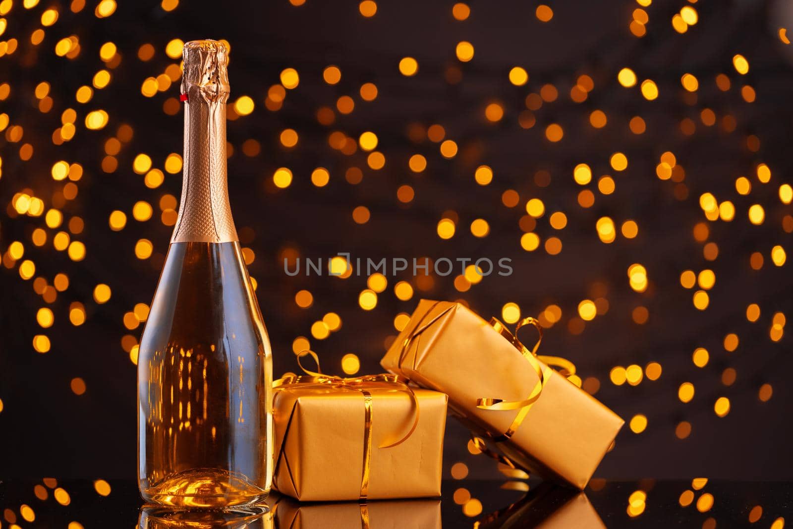 Champagne bottle and wrapped gift on blurred background of Christmas lights front view