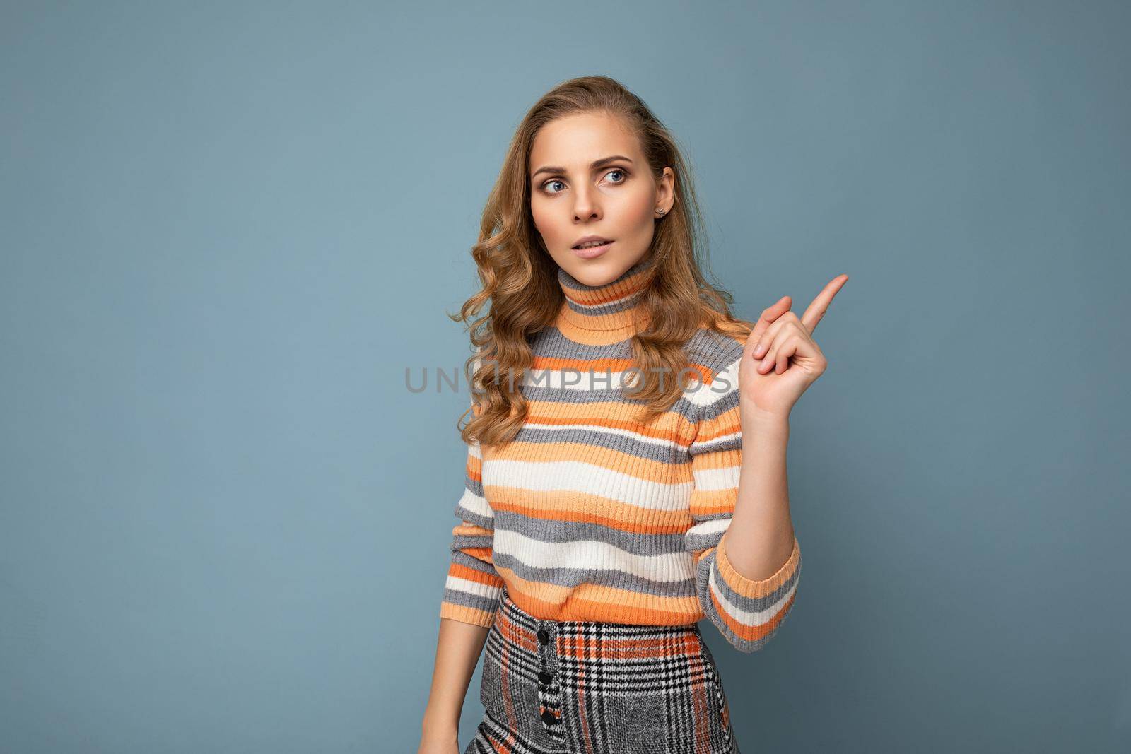 Photo portrait of young pretty beautiful concentrated serious blonde woman wearing stylish colourful striped pullover isolated on blue background with copy space and shows finger, having doubts. Pay attention concept.