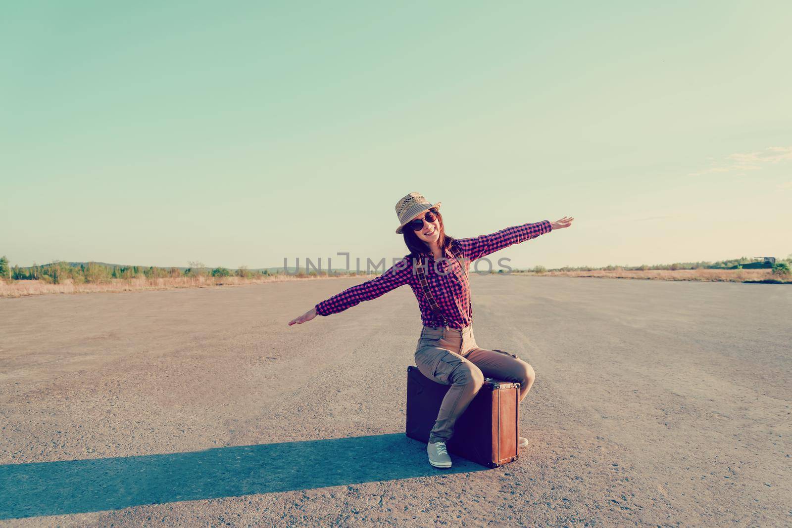Happy traveler woman sits on vintage suitcase on road and makes a gesture of flight