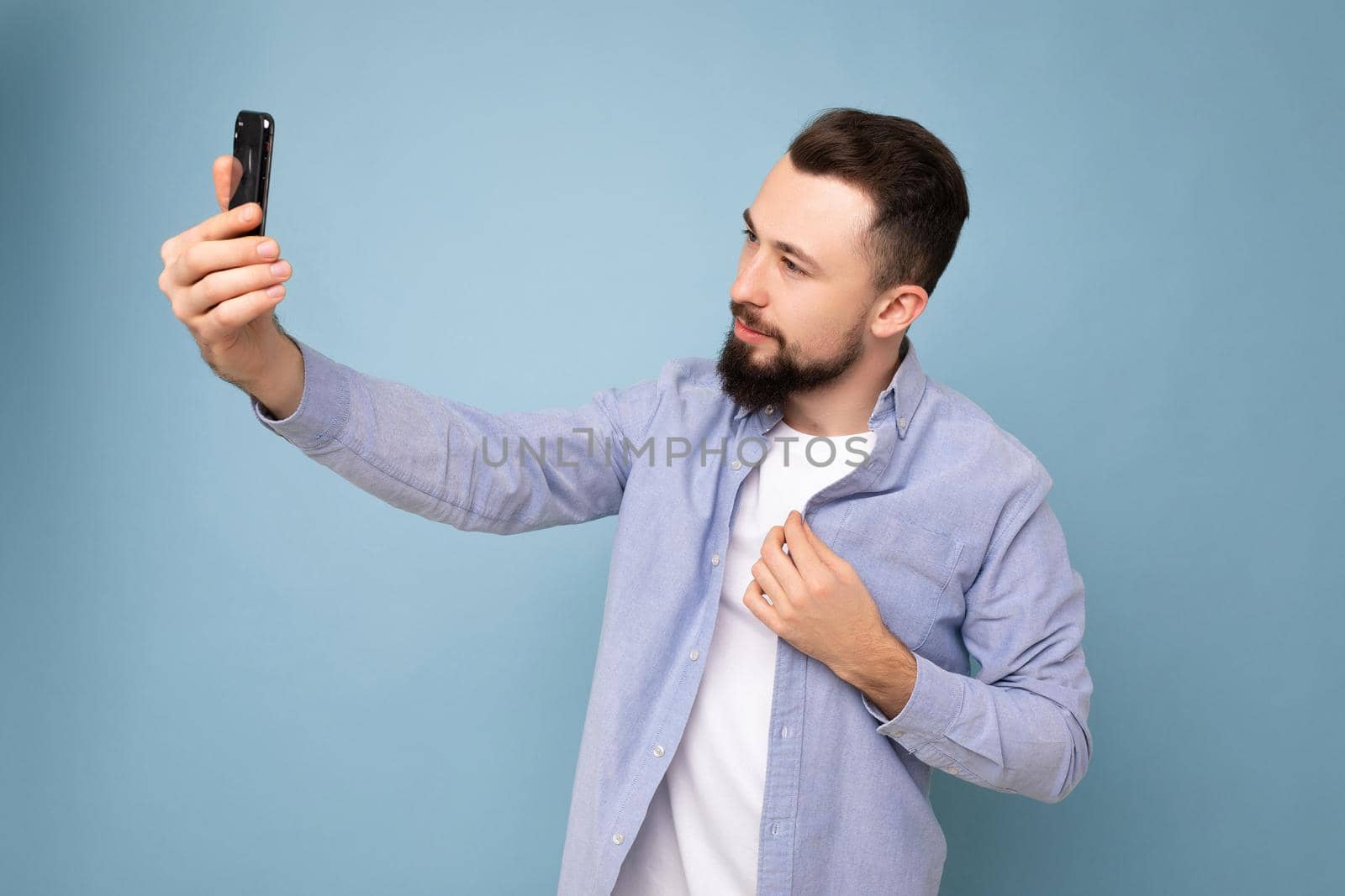 Handsome young man wearing casual stylish clothes standing isolated over background wall holding smartphone taking selfie photo looking at mobile phone screen display.