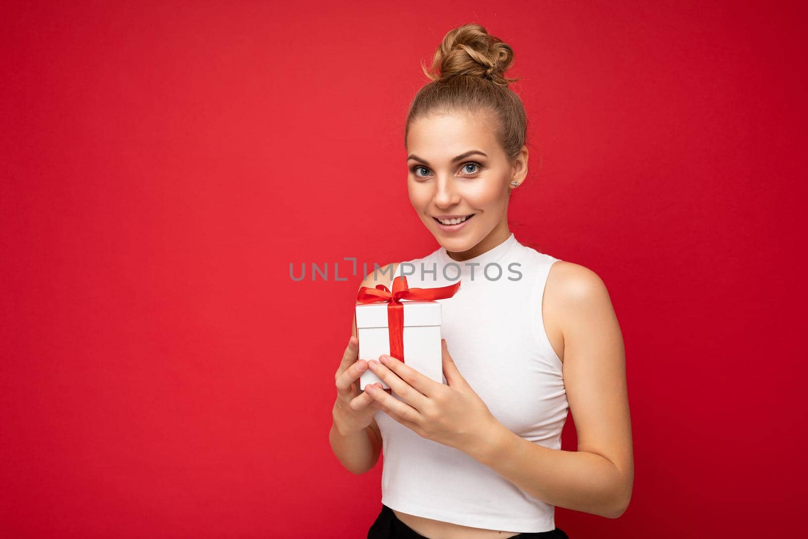 Shot of beautiful positive smiling young blonde woman isolated over red background wall wearing white top holding gift box and looking at camera. Copy space, mockup