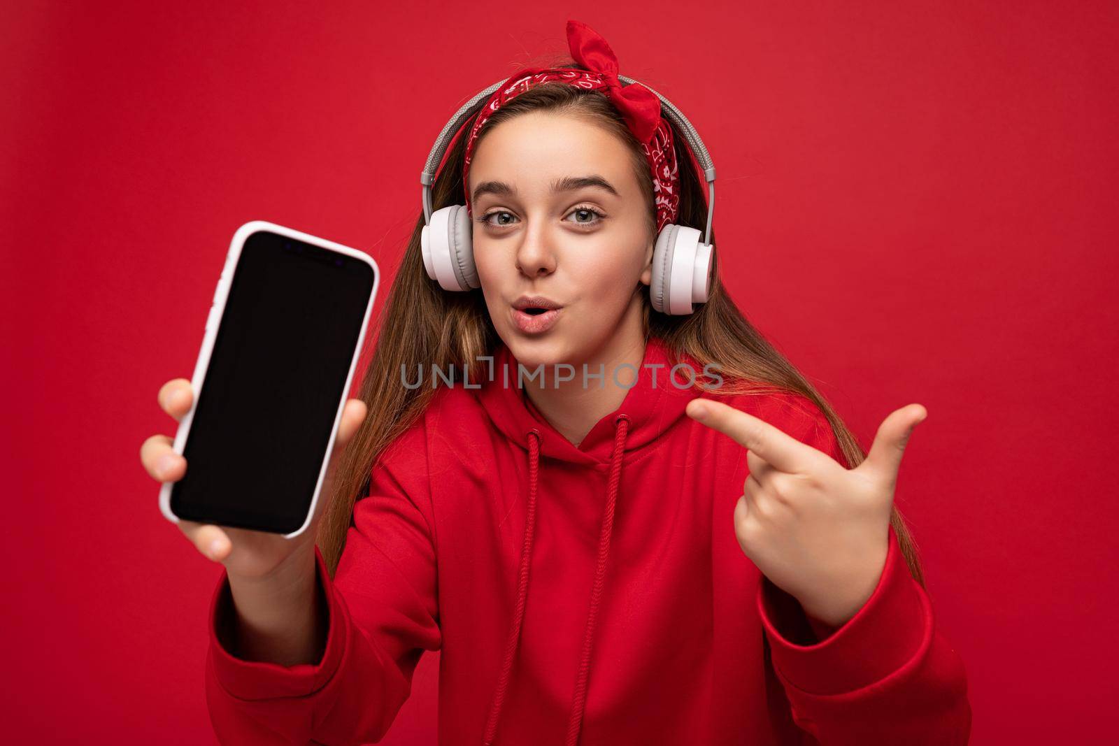 Attractive joyful amazed brunette girl wearing red hoodie isolated on red background holding and showing smartphone with empty display for cutout wearing white wireless headphones listening to funny music looking at camera and pointing finger at device by TRMK