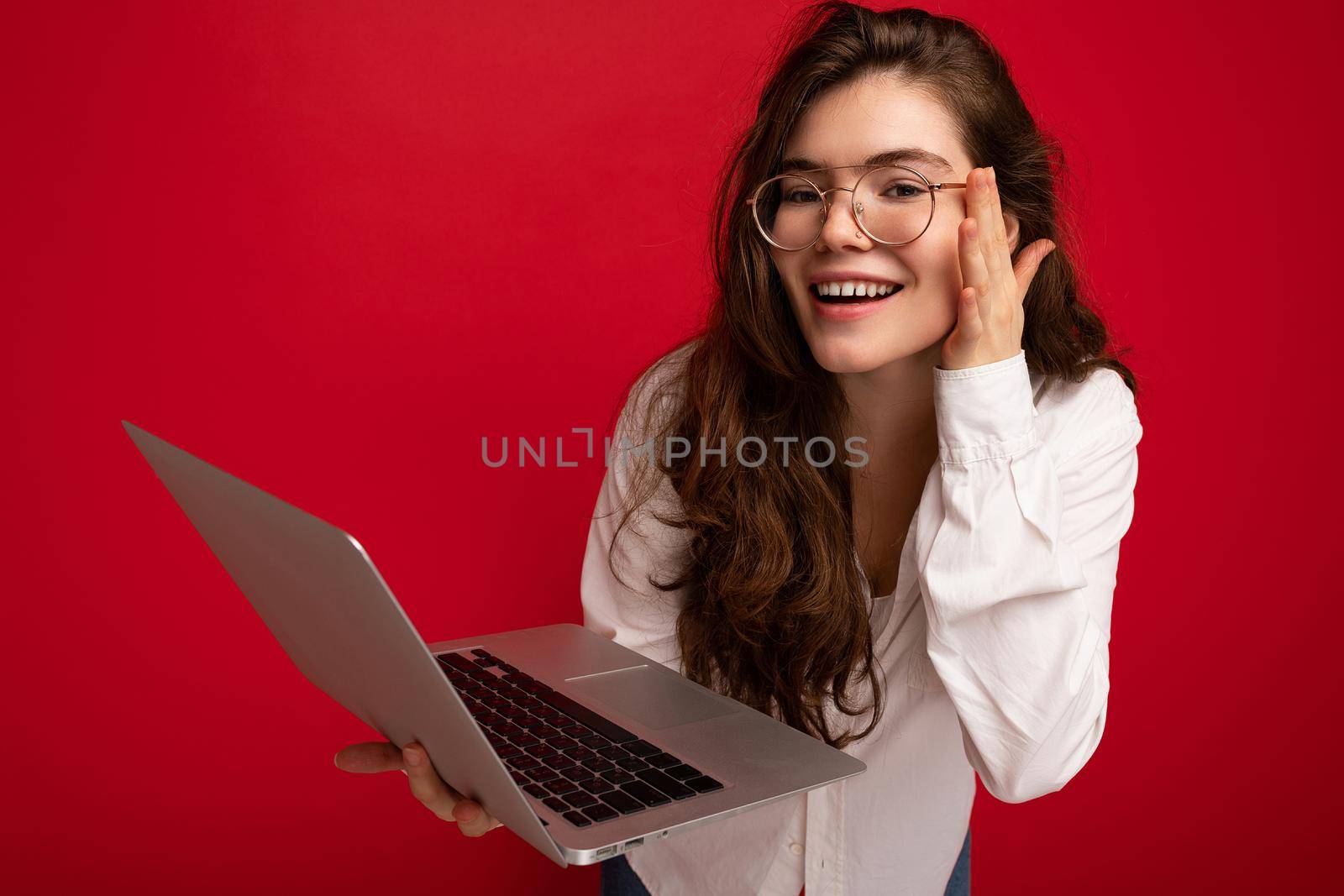 Side profile Photo shot of beautiful smiling brunet curly young woman holding computer laptop wearing glasses white shirt looking at netbook display isolated over red wall background.