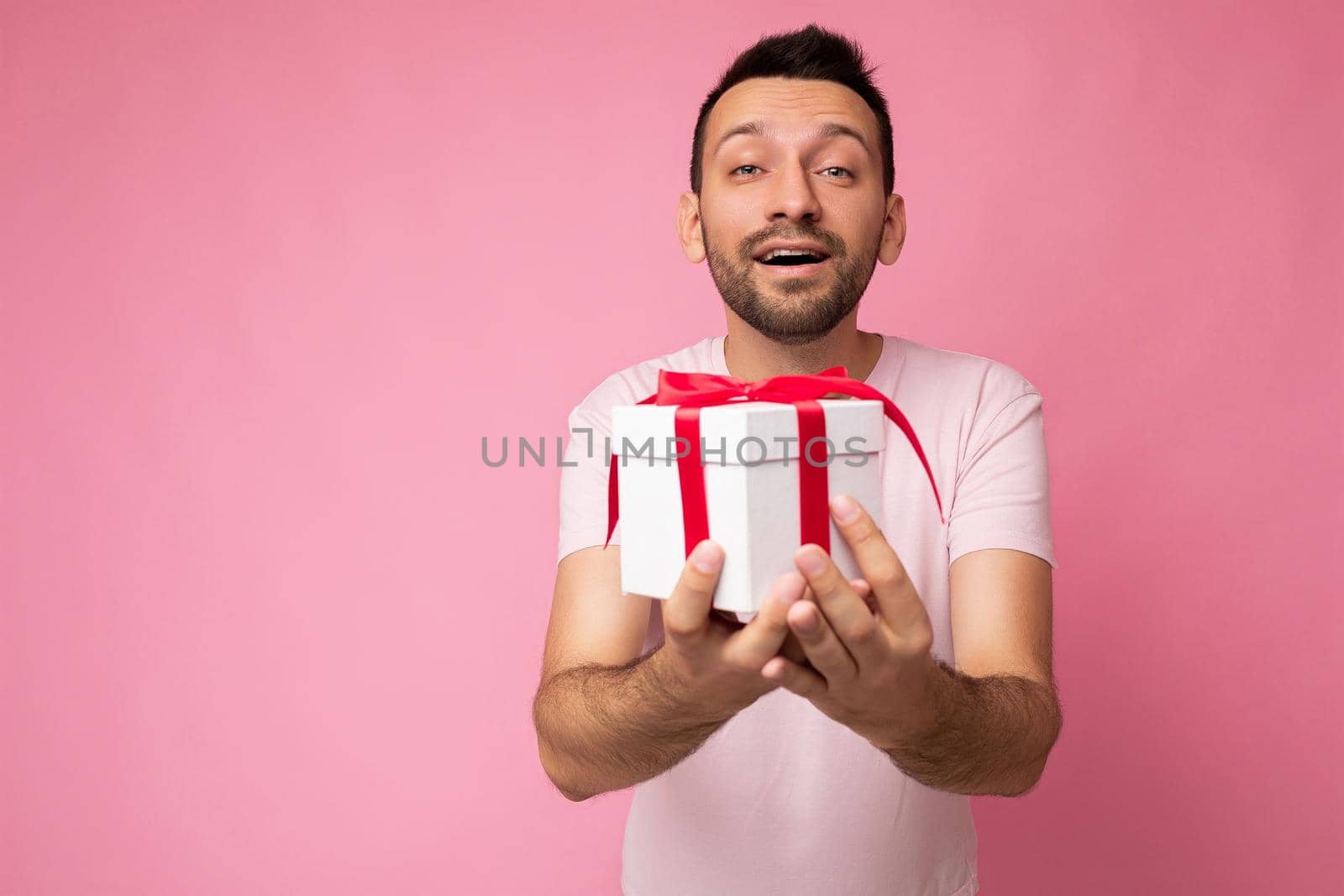 Photo shot of handsome positive happy kind brunette young unshaven man with beard isolated over pink background wall wearing casual pink t-shirt holding white gift box with red ribbon and looking at camera by TRMK