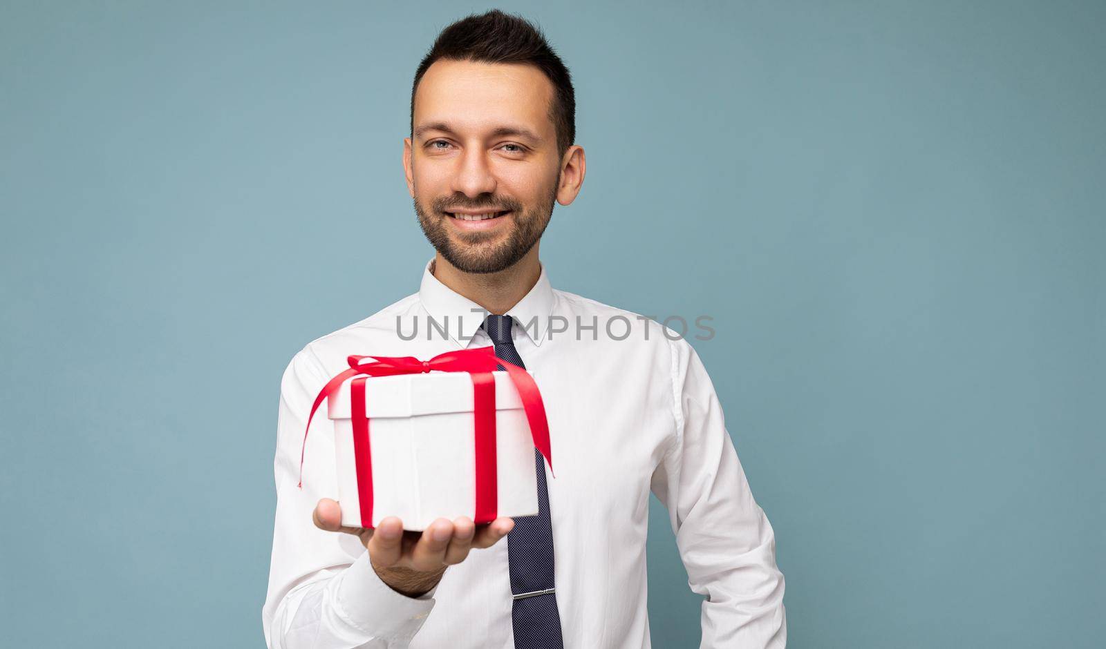 Photo shot of handsome happy smiling brunet young unshaven man isolated over blue background wall wearing white shirt and tie holding white gift box with red ribbon and looking at camera.