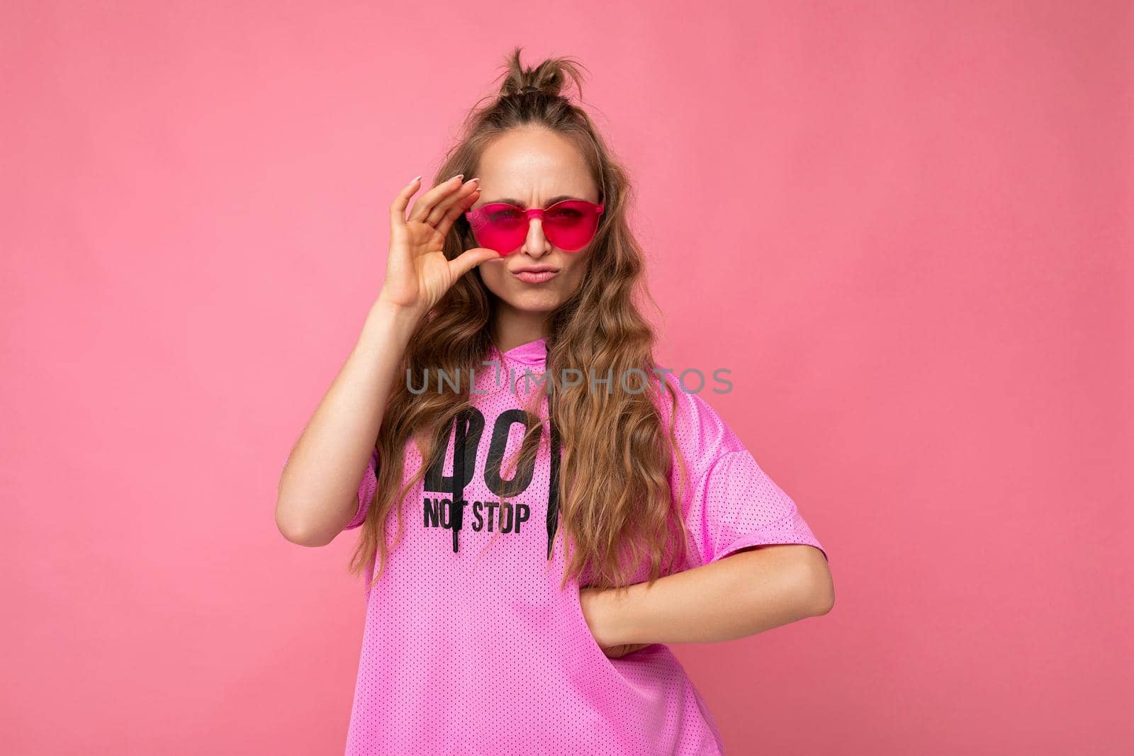Photo shot of pretty sad dissatisfied young blonde curly woman isolated over pink background wall wearing casual pink t-shirt and stylish colourful sunglasses looking at camera and having questions.