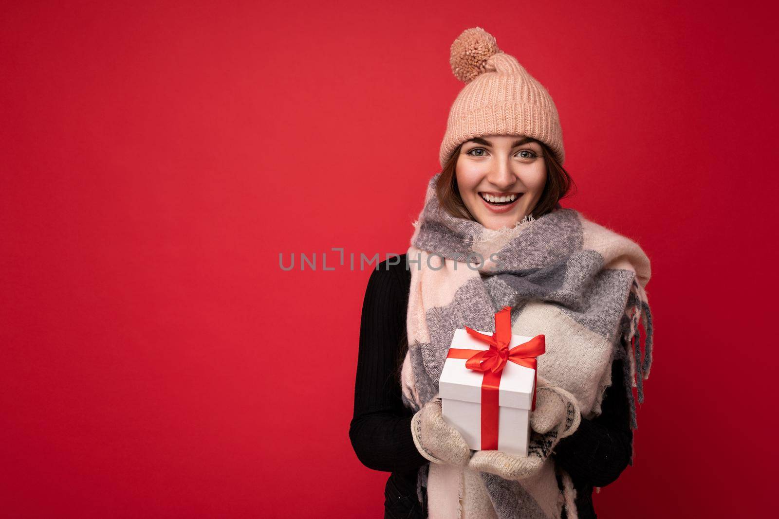 Beautiful happy young brunette woman isolated over colourful background wall wearing stylish casual clothes holding gift box and looking at camera. Free space