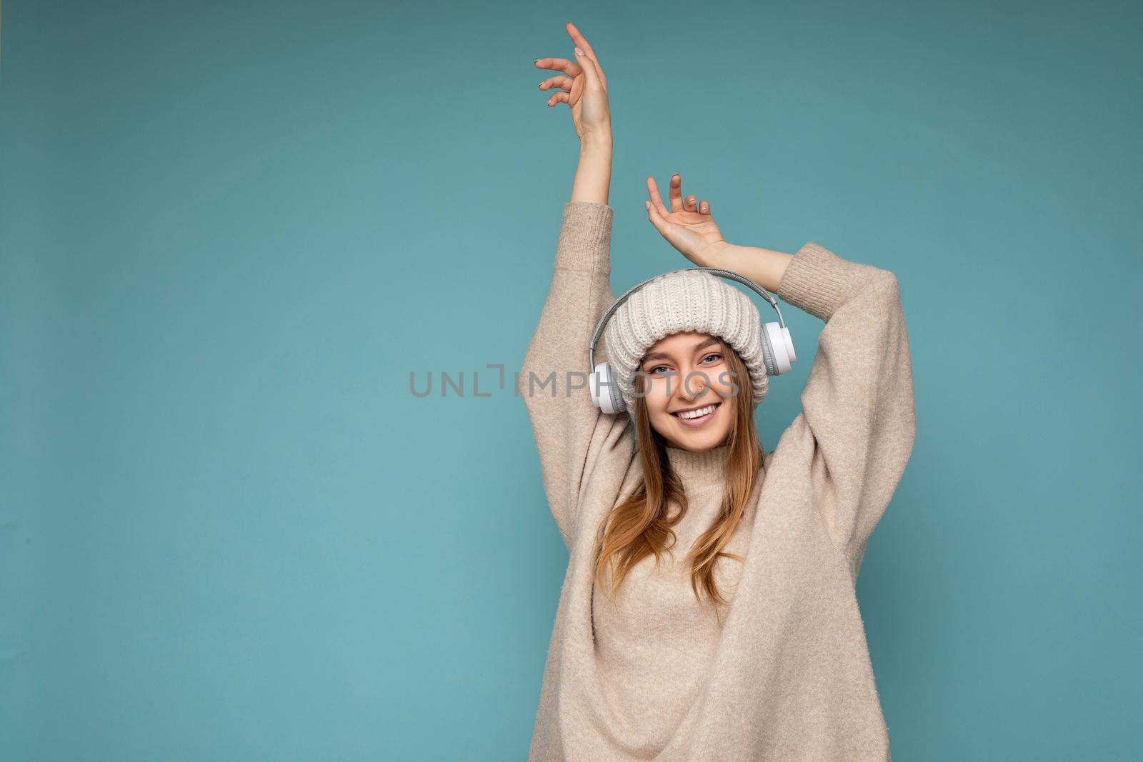 Attractive positive smiling young blonde woman wearing beige winter sweater and hat isolated over blue background wall wearing white wireless bluetooth earphones listening to music having fun and looking at camera. free space