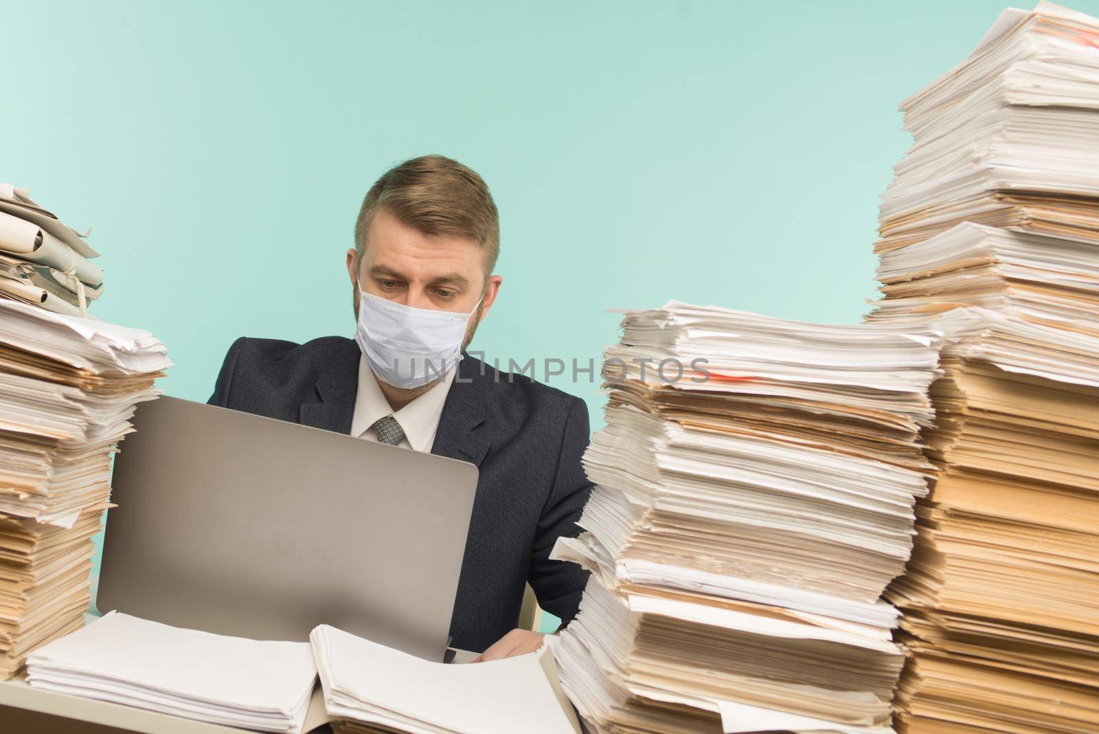 A male accountant or company manager works in an office in a pandemic in view of the accumulated paper work. A protective medical mask is on the face. On the desktop are large stacks of documents. by zartarn