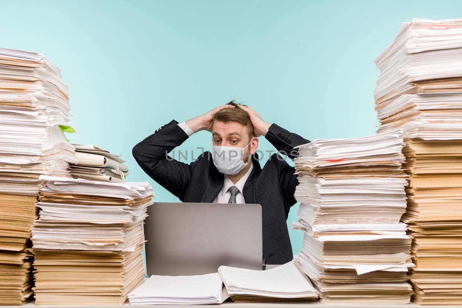 A male accountant or company manager works in an office in a pandemic in view of the accumulated paper work. A protective medical mask is on the face. On the desktop are large stacks of documents. - image