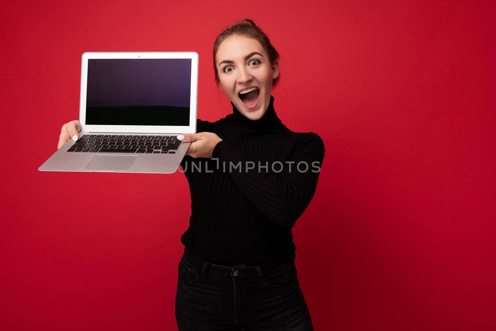 Close-up portrait of Beautiful smiling happy young woman with gathered dark hair holding computer laptop with empty monitor screen with mock up wearing black longsleeve looking at camera.