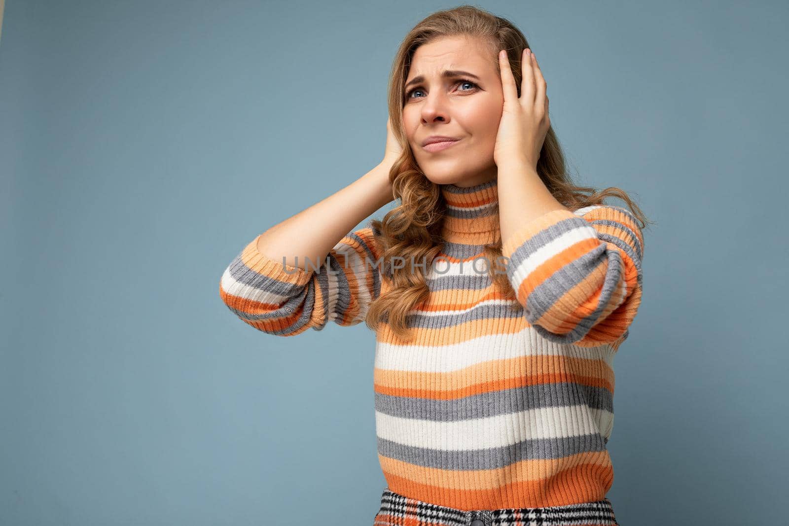 Photo of young dissatisfied attractive blonde curly woman with sincere emotions wearing casual striped pullover isolated on blue background with copy space and covering ears trying not to hear.