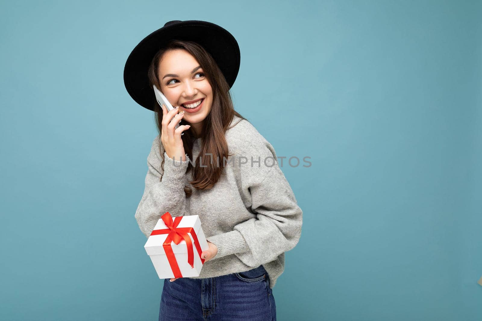 Photo of beautiful happy positive joyful young brunette woman isolated over blue background wall wearing black hat and grey sweater holding gift box talking on mobile phone and looking to the side. Copy space, mockup