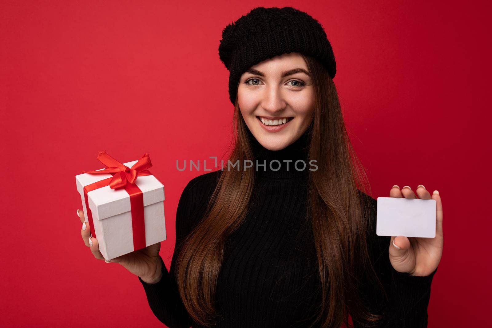 Closeup portrait Attractive positive young brunette woman wearing black sweater and hat isolated on red background holding credit card and gift box looking at camera.