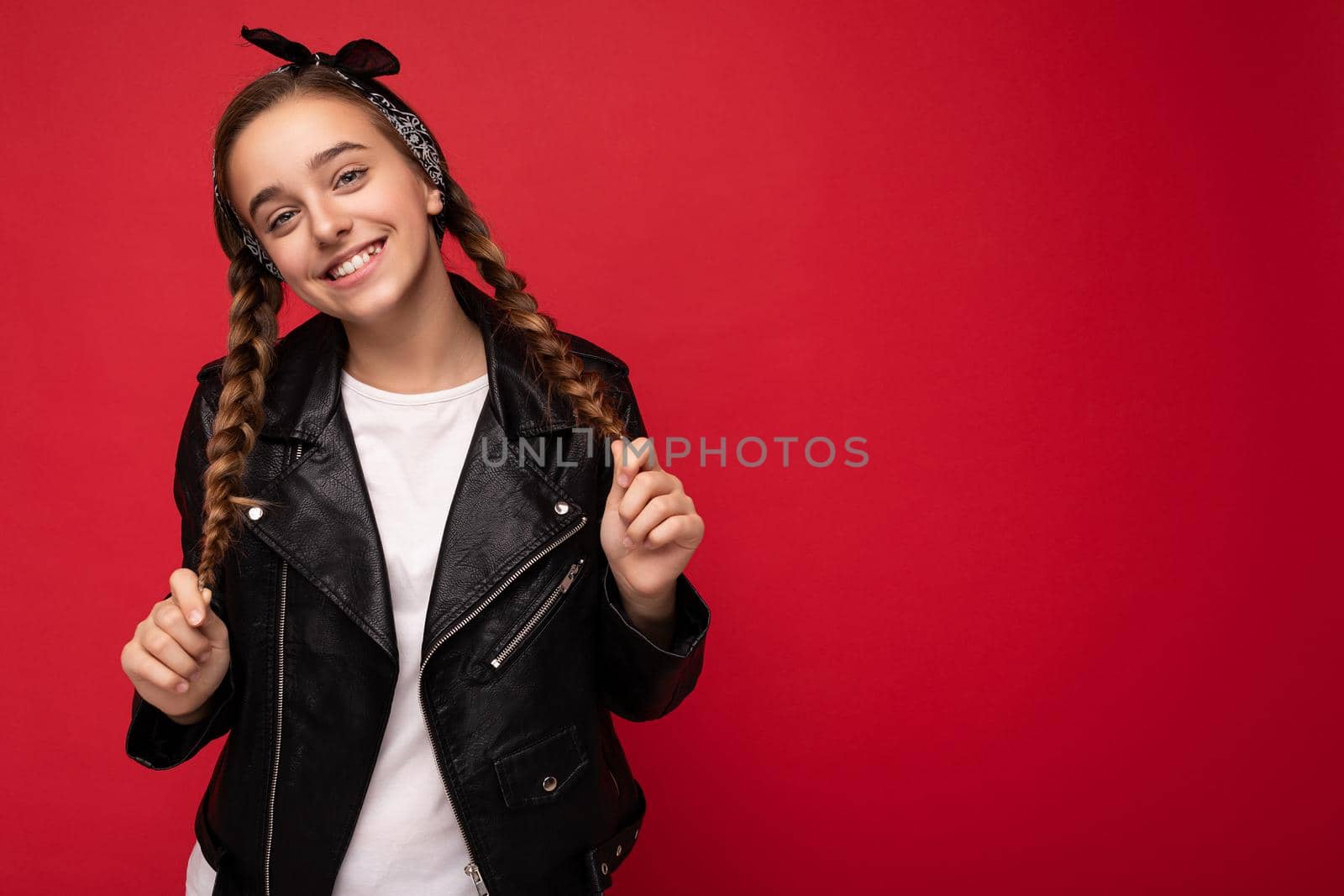 Photo shot of pretty positive smiling brunette little female teenager with pigtails wearing stylish black leather jacket and white t-shirt standing isolated over red background wall looking at camera. Copy space