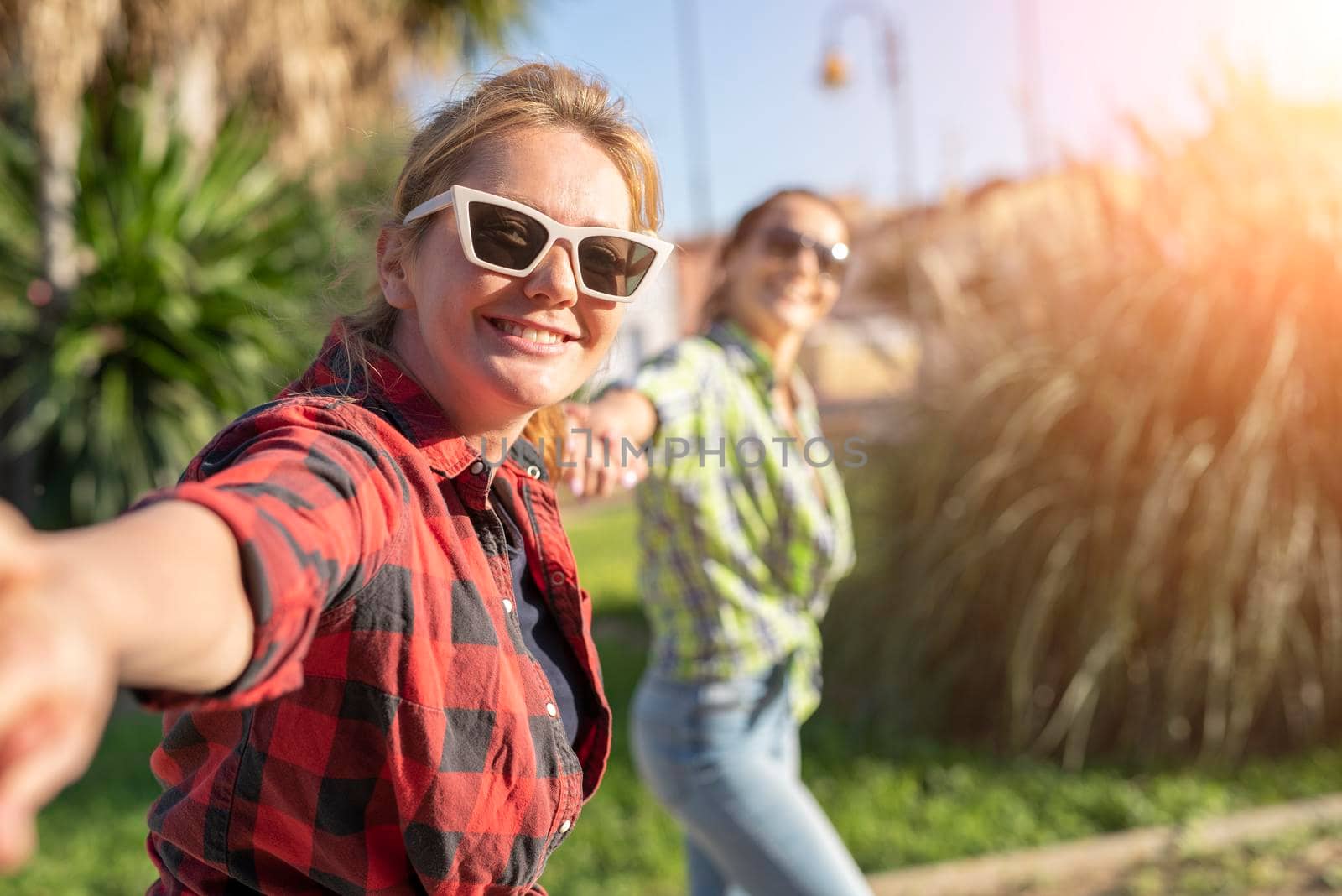 Pretty young caucasian woman in sunglasses pulling along by the hand invitingly with a happy smile as her friend on the background. Sunny summer day.