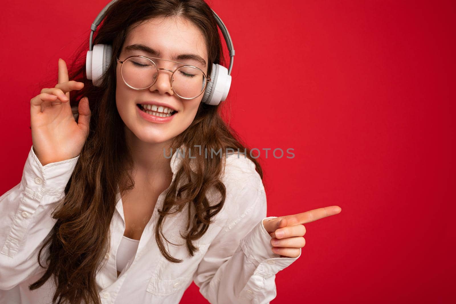 Closeup photo of attractive positive smiling young brunet woman wearing white shirt and optical glasses isolated over red background wearing white wireless bluetooth headsets listening to music and moving having fun. free space