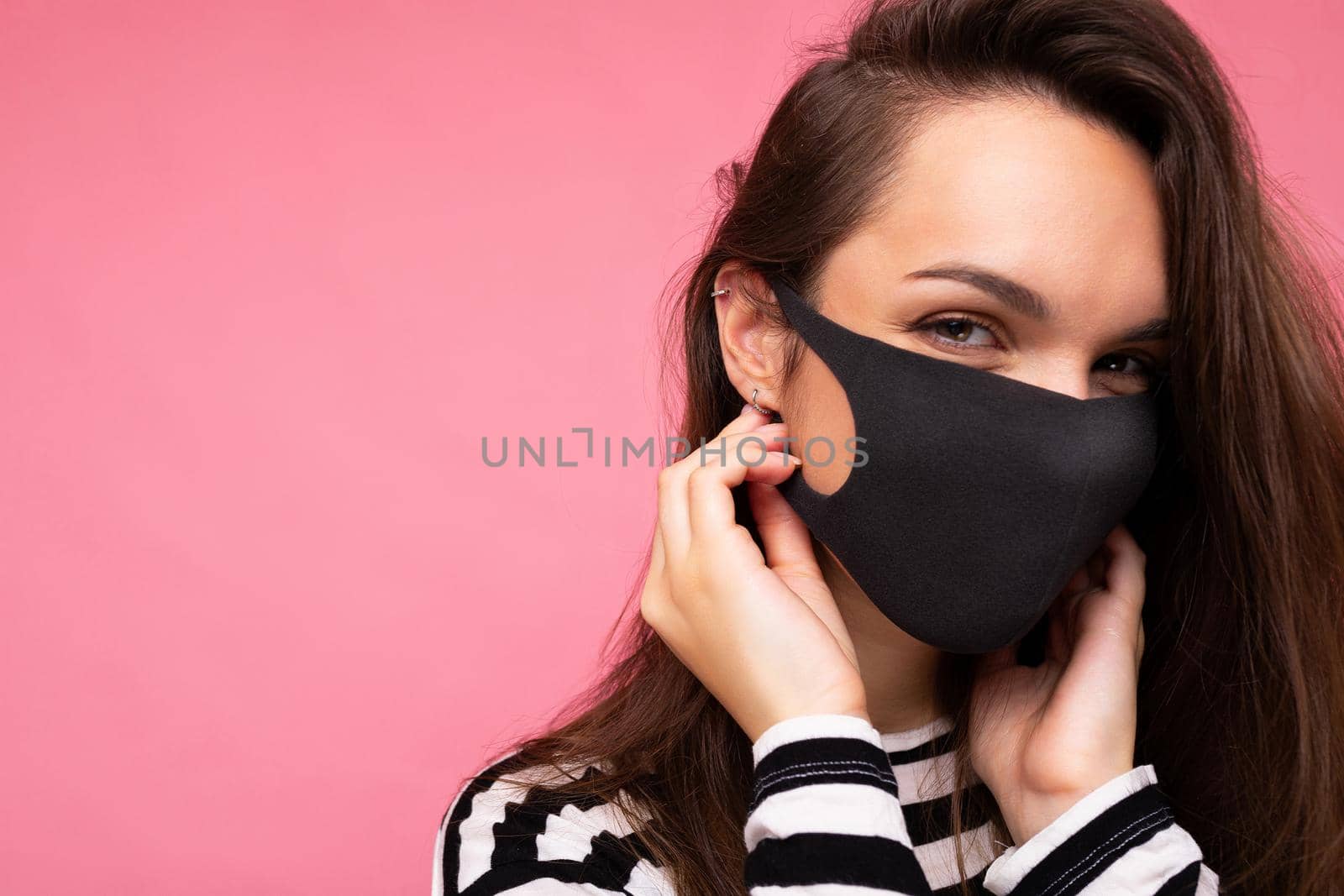 Woman wearing stylish protective face mask, posing on pink background. Trendy fashion accessory during quarantine of coronavirus pandemic. Close up studio portrait. Copy, empty space for text.
