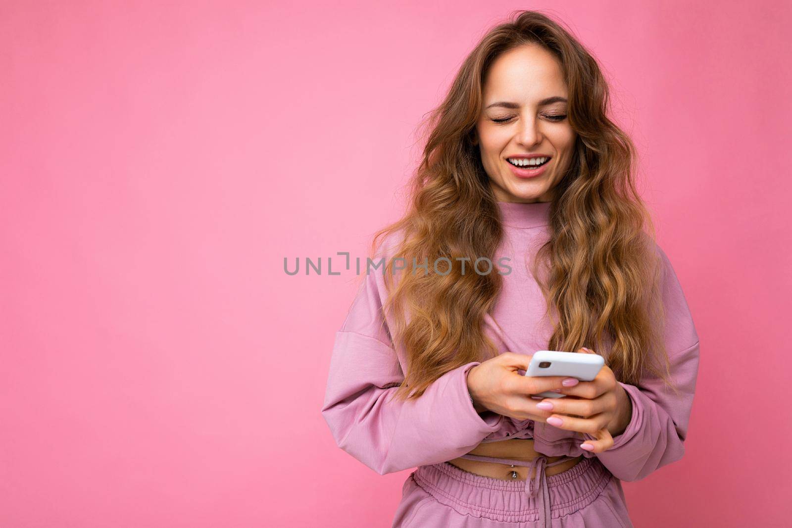 Photo shot of winling attractive positive good looking young woman wearing casual stylish outfit poising isolated on background with empty space holding in hand and using mobile phone messaging sms looking at camera.