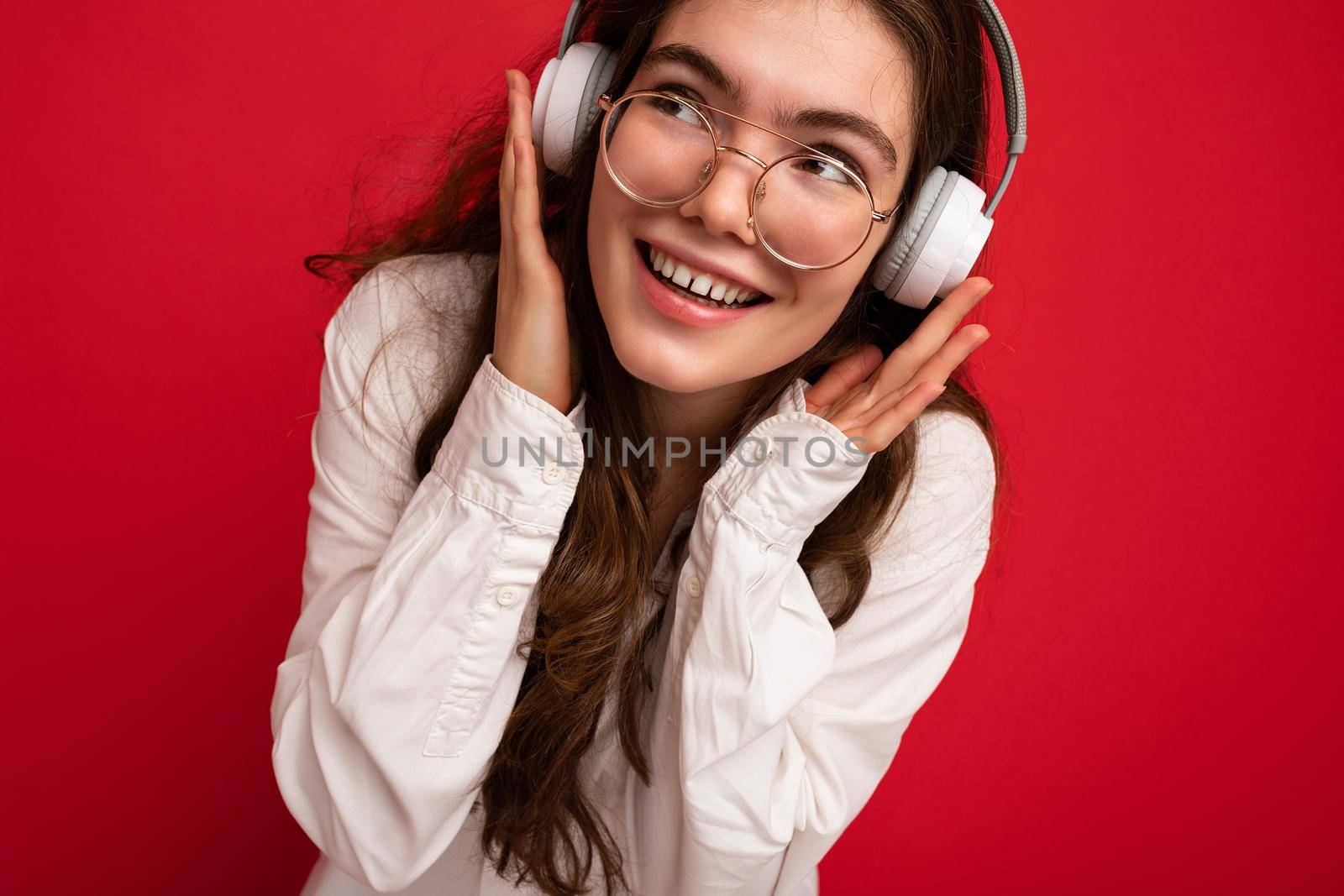 Closeup photo of attractive positive smiling young brunet woman wearing white shirt and optical glasses isolated over red background wearing white wireless bluetooth headsets listening to music and having fun looking to the side.