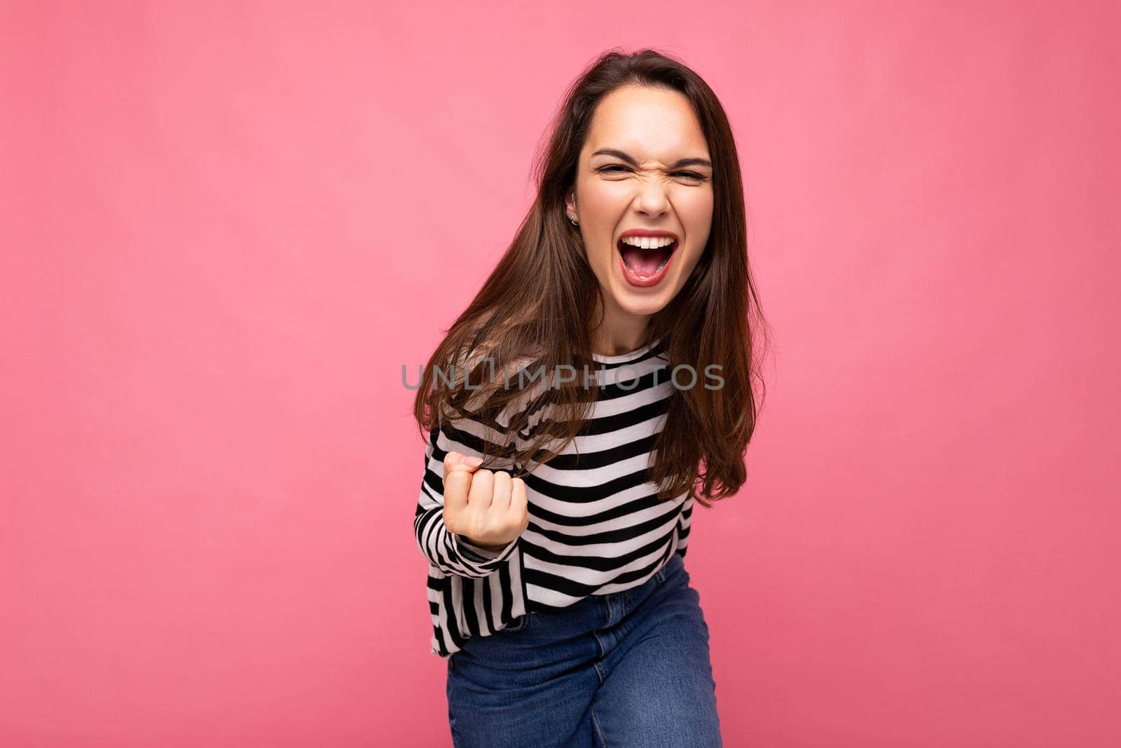 Portrait of young positive happy beautiful brunet female person with sincere emotions wearing casual striped pullover isolated on pink background with empty space and celebrating winning shouting yes.