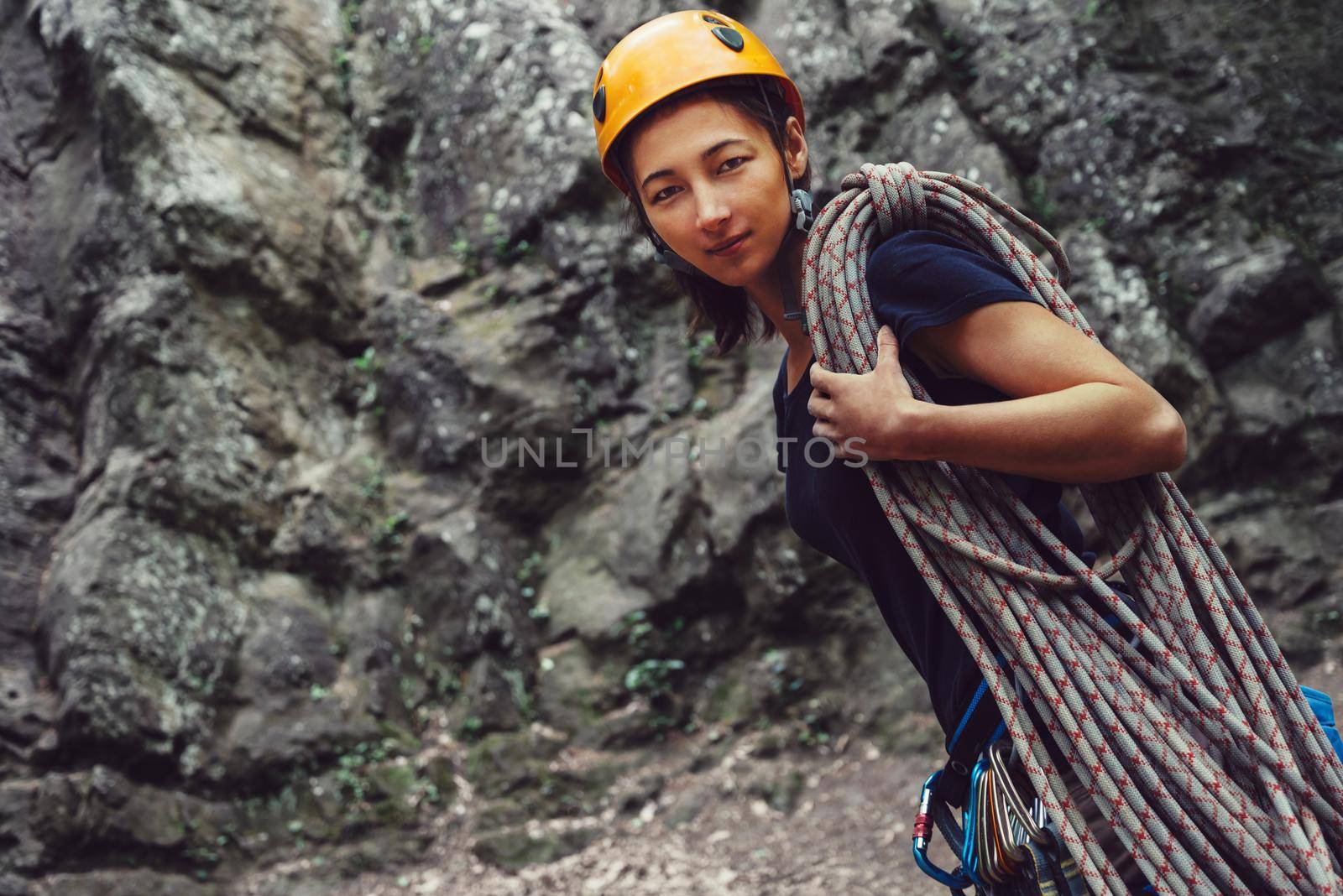 Smiling young woman standing with climbing equipment and rope in front of a stone rock outdoor