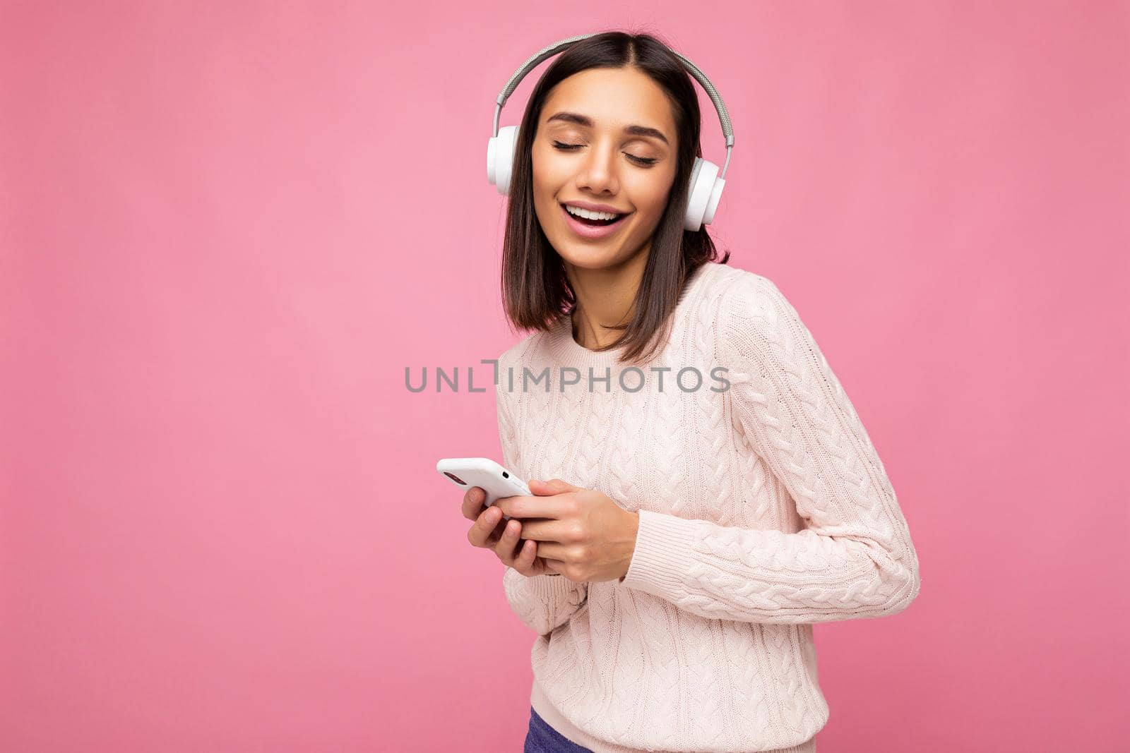 Photo shot of beautiful joyful smiling young female person wearing stylish casual outfit isolated over colorful background wall wearing white bluetooth wireless earphones and listening to music and using mobile phone with close eyes. copy space