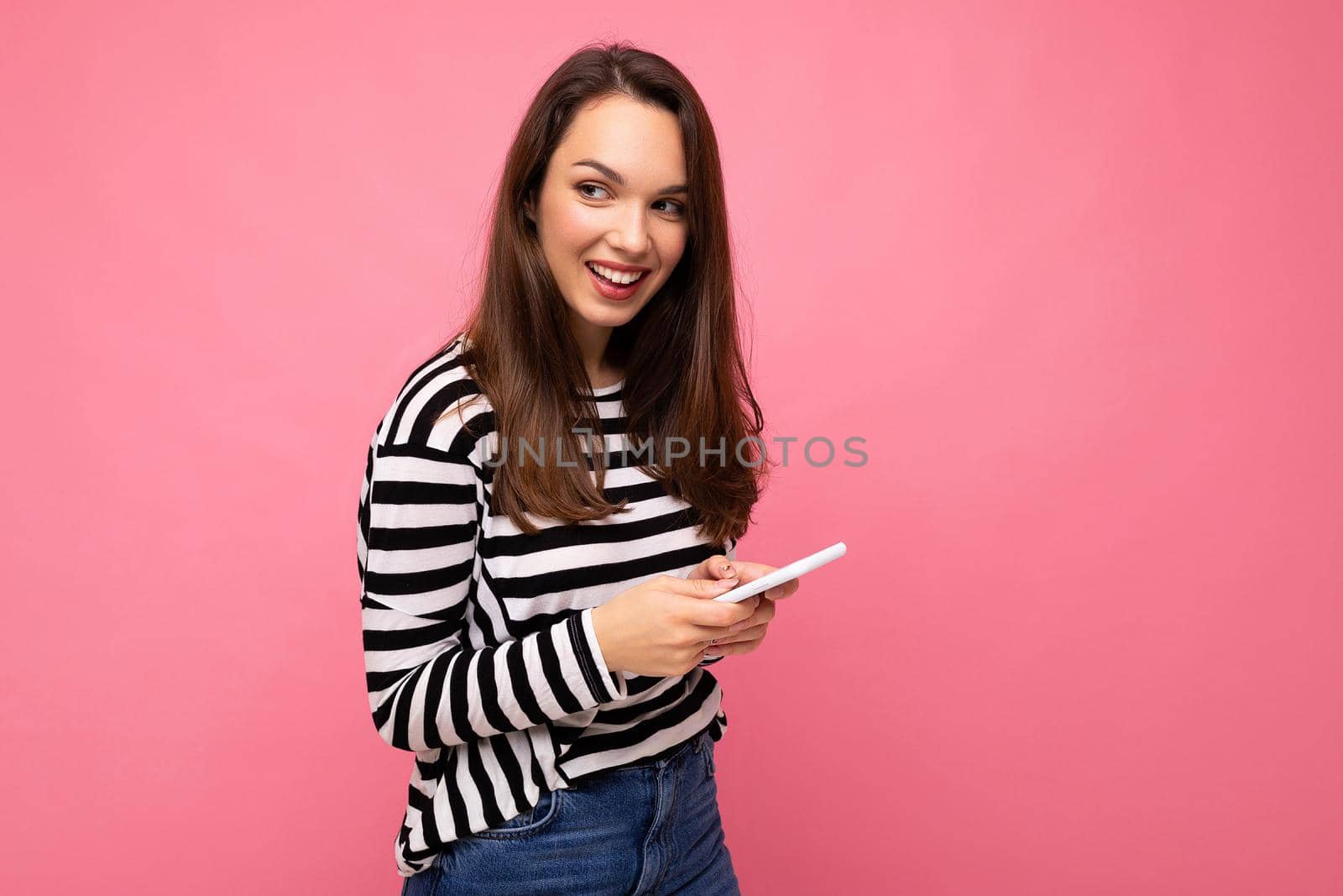 side profile shot of pretty young brunette woman using mobile phone communicating via texting sms wearing sweater isolated on wall background looking to the side.