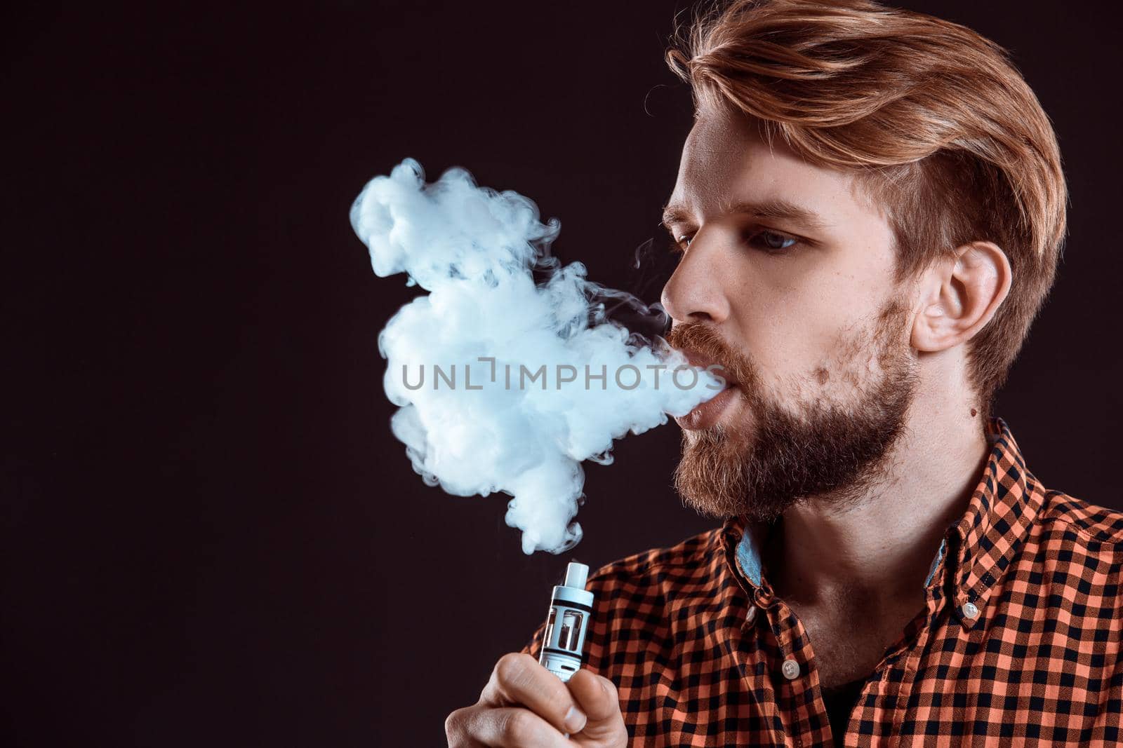 young man wearing a plaid shirt smokes an electronic cigarette on a black background. close-up