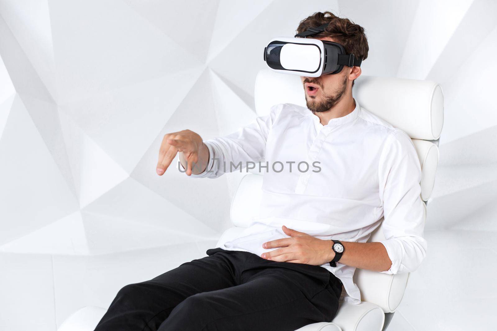 Excited young man is getting experience using VR-headset glasses of virtual reality gesticulating with his hands. A young man sits on a comfortable armchair in a room with white walls