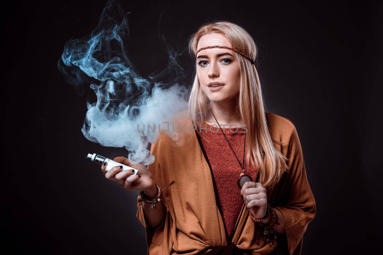 Young woman in the Boho style blowing smoke. The blonde on a dark background. Young woman looking into the camera