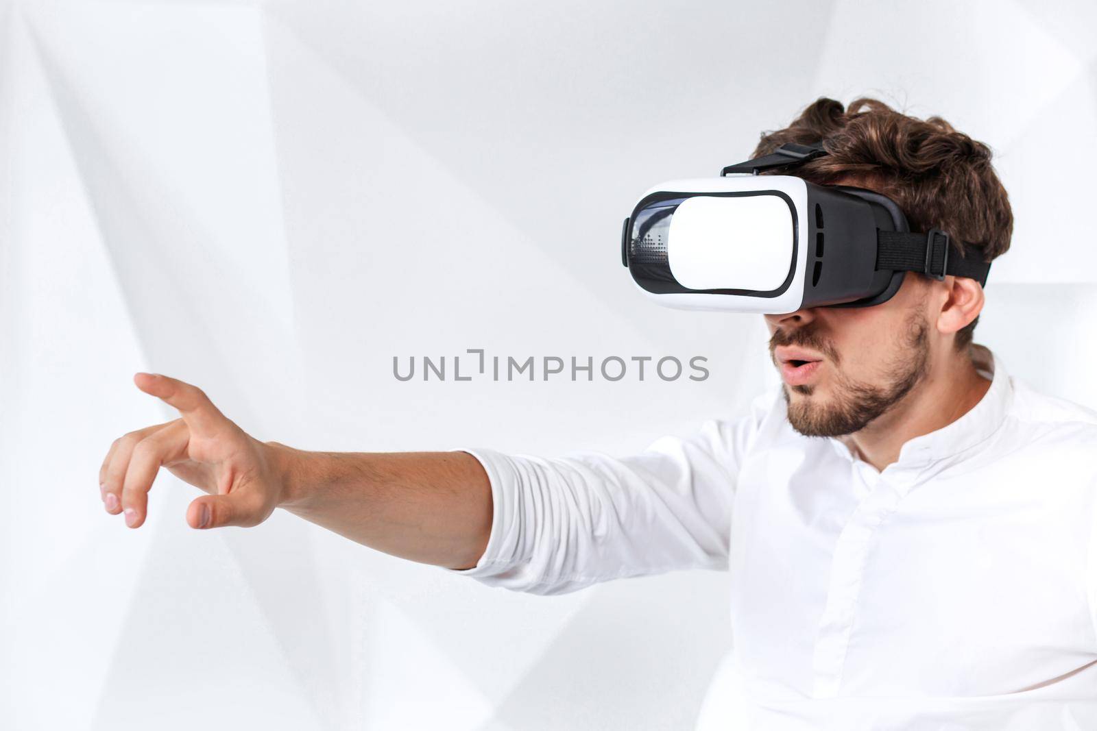 Excited young man is getting experience using VR-headset glasses of virtual reality gesticulating with his hands. A young man sits on a comfortable armchair in a room with white walls