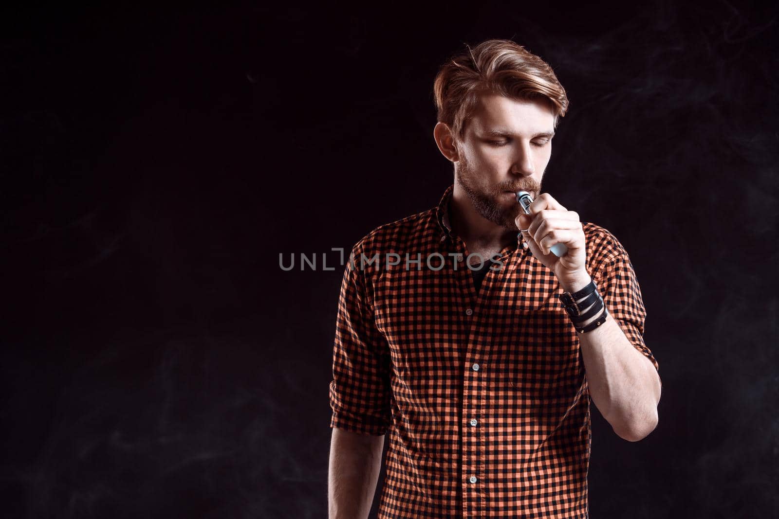 young man wearing a plaid shirt smokes an electronic cigarette on a black background