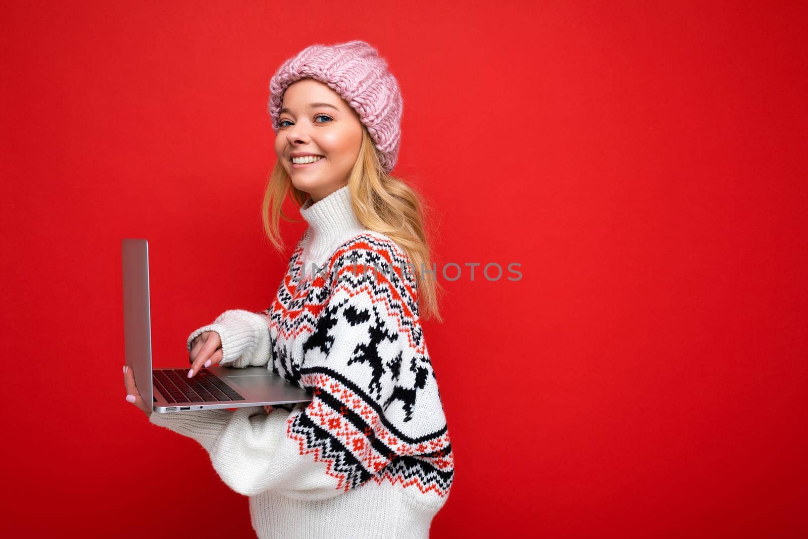 side profile photo of charming smiling happy pretty young woman holding laptop isolated over red wall background looking camera wearing winter hat and knitted new year sweater typing on keyboard.