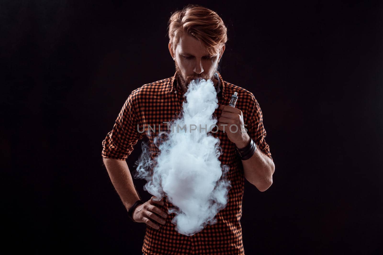 young man wearing a plaid shirt smokes an electronic cigarette on a black background