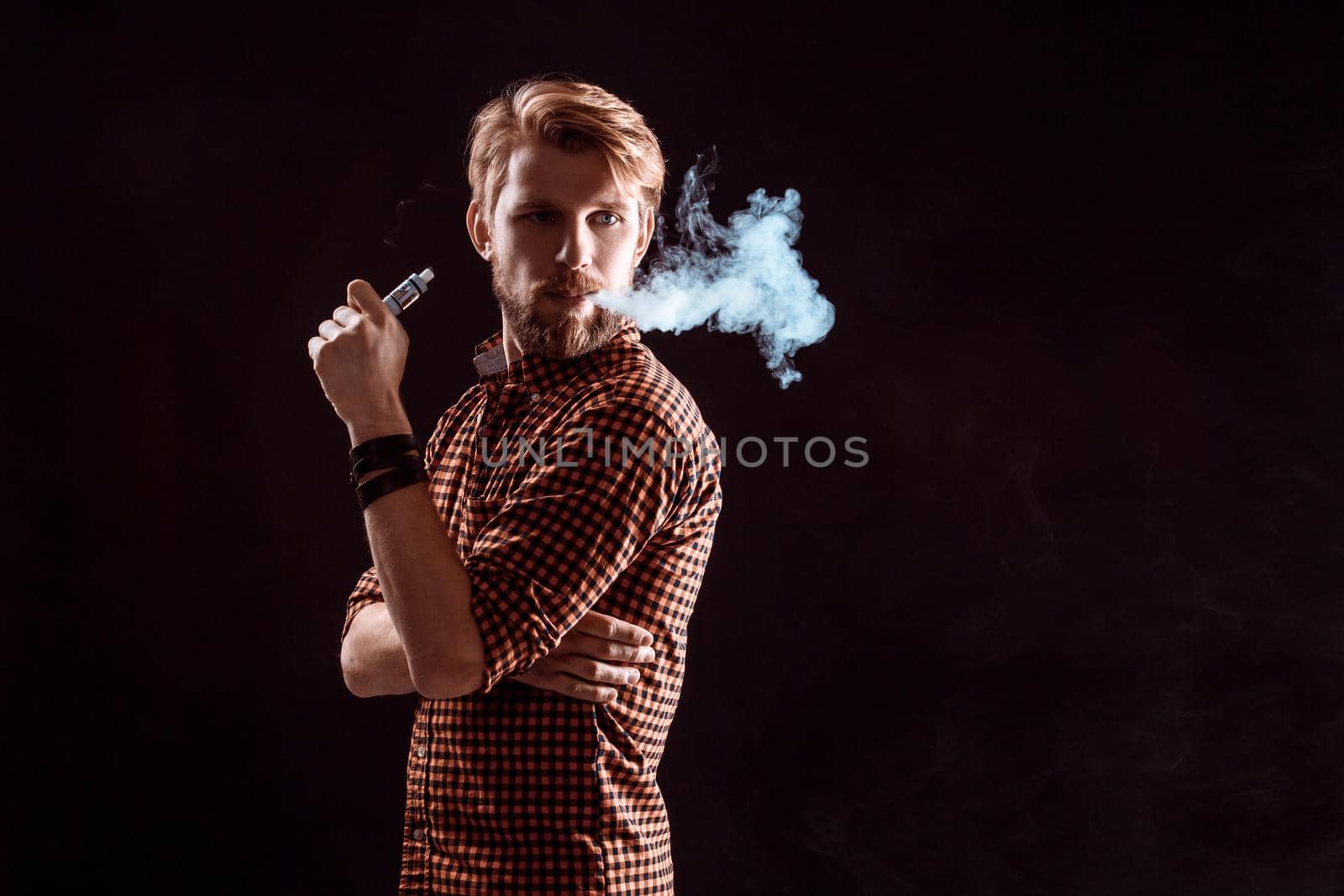 young man wearing a plaid shirt smokes an electronic cigarette on a black background