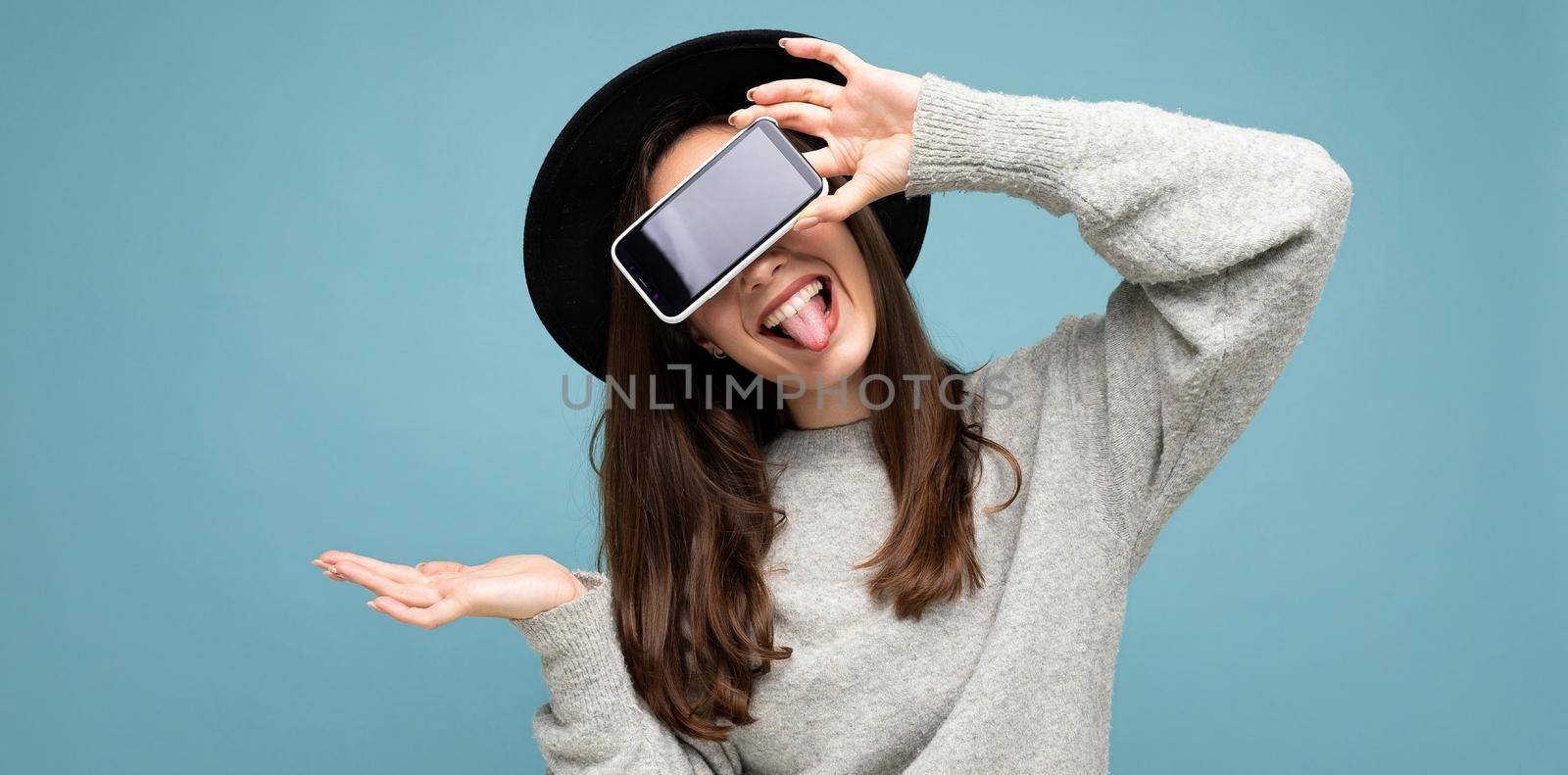 beautiful young woman wearing black hat and grey sweater holding phone showing smartphone showing tongue isolated on background.Mock up, cutout, empty space