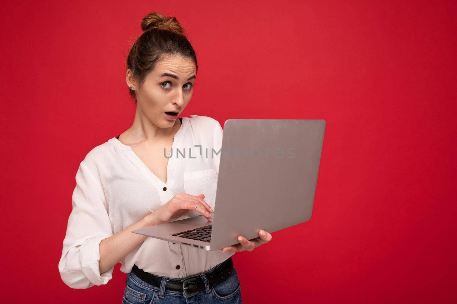 Side profile of Beautiful surprised brunet young woman holding netbook computer looking at camera wearing white shirt typing on keyboard isolated on red background.