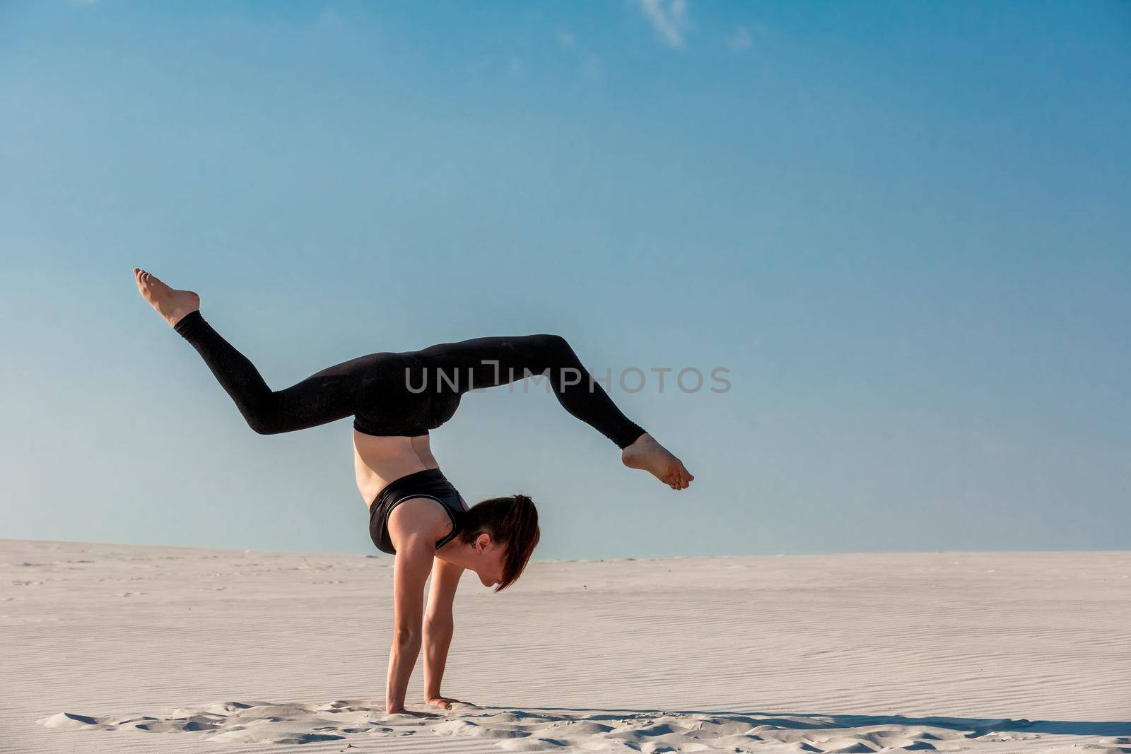 Young woman practicing inversion balancing yoga pose handstand on beach with white sand and bright blue sky