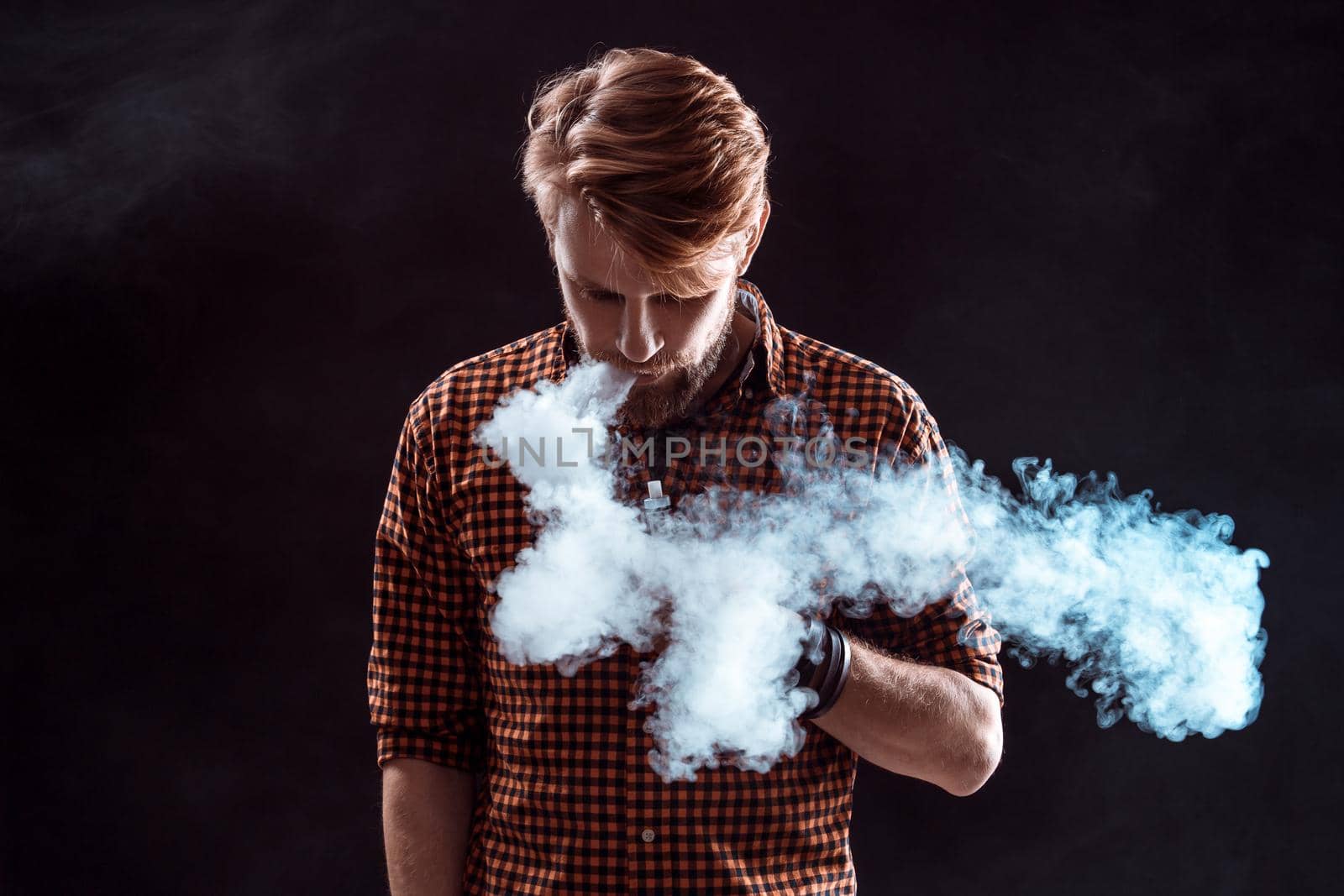 young man wearing a plaid shirt smokes an electronic cigarette on a black background