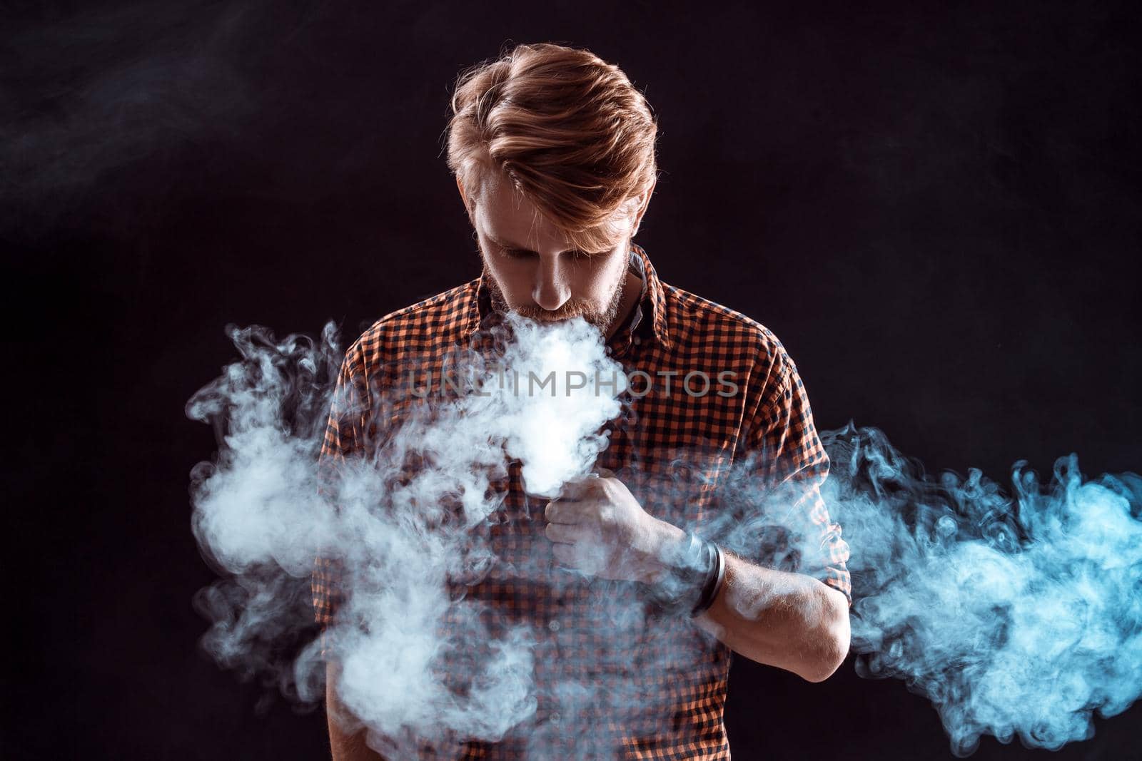 young man wearing a plaid shirt smokes an electronic cigarette on a black background