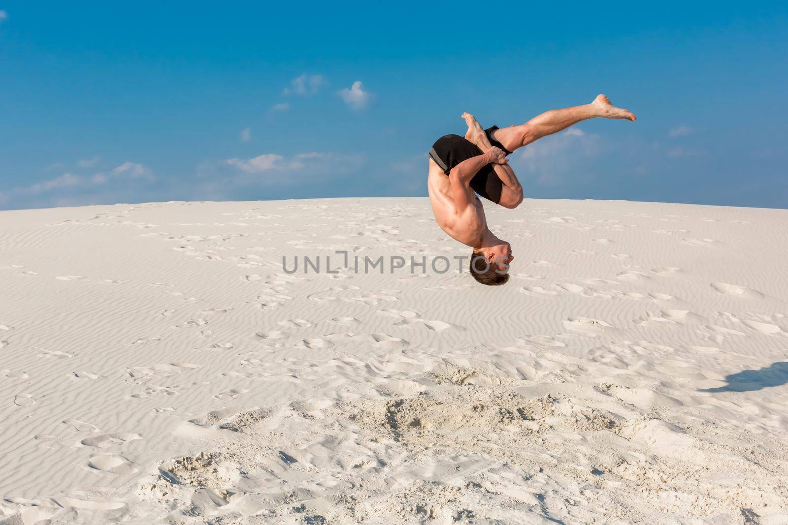 Portrait of young parkour man doing flip or somersault on the sand. Freezed moment of beginning doing bounce.