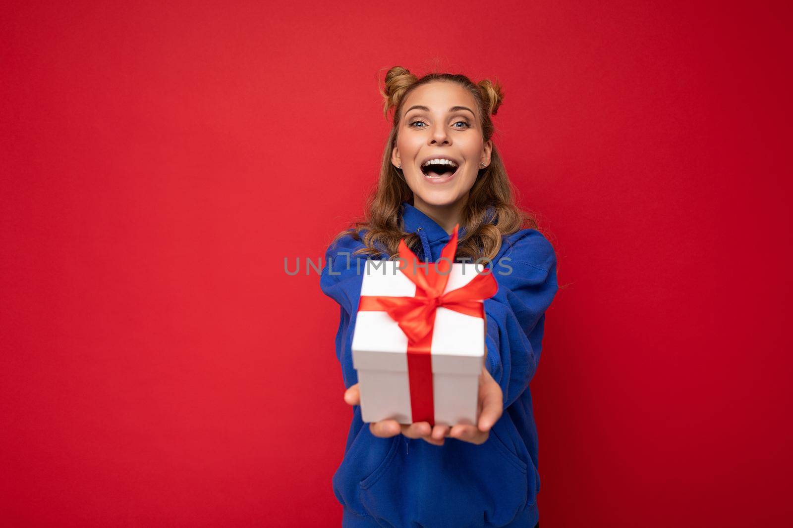Photo shot of pretty positive surprised amazed young blond woman isolated over colourful background wall wearing trendy outfit look holding gift box and looking at camera.