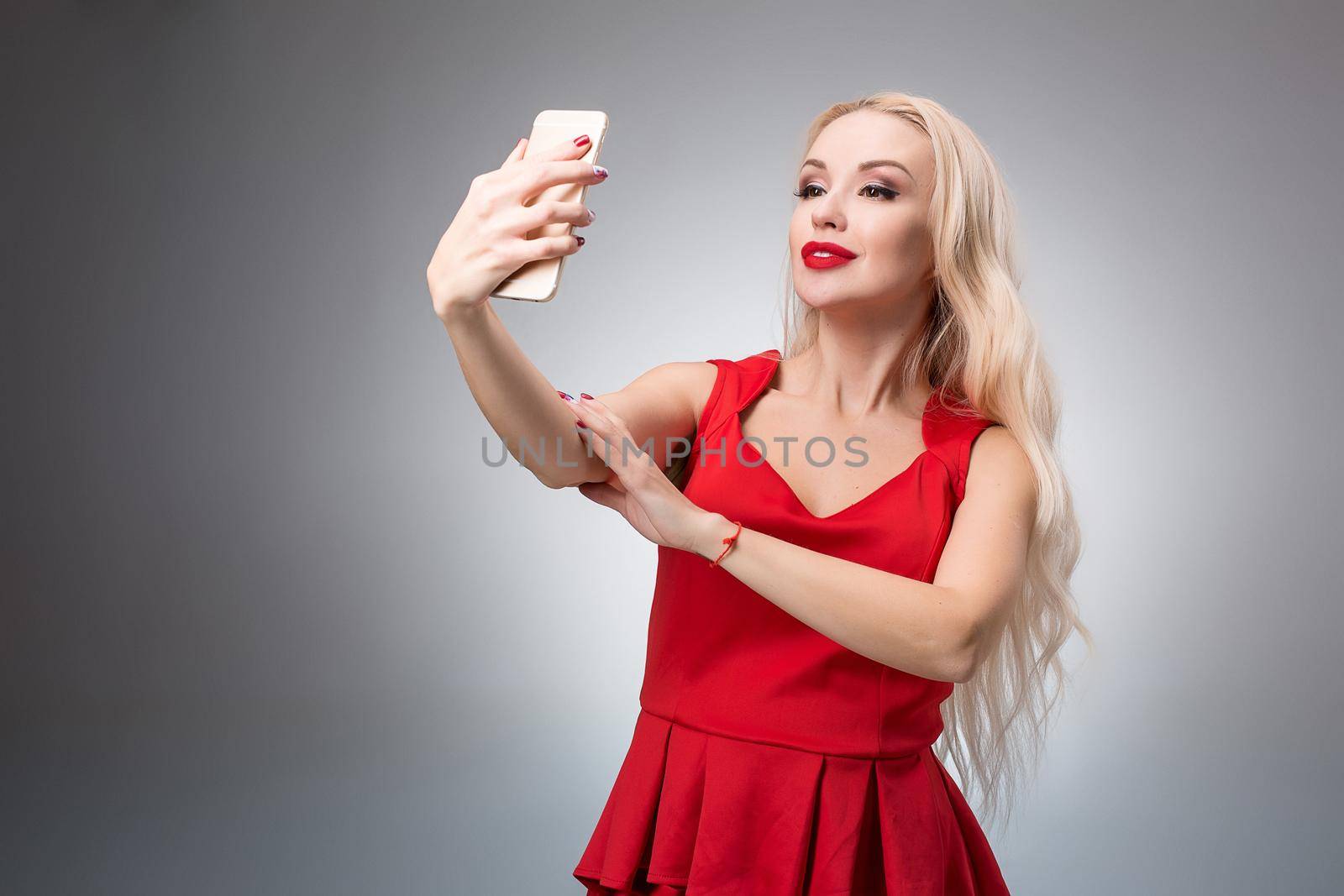 Portrait of a Beautiful successful smiling girl doing selfie in red dress on light background by nazarovsergey
