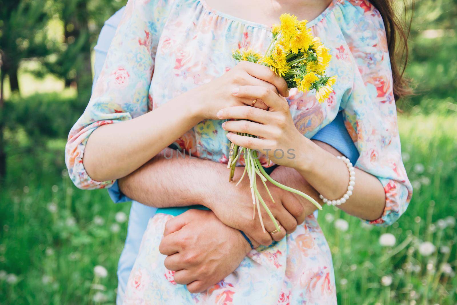 Married loving couple resting in summer, man embraces a woman in summer park. Woman holding bouquet of yellow dandelions