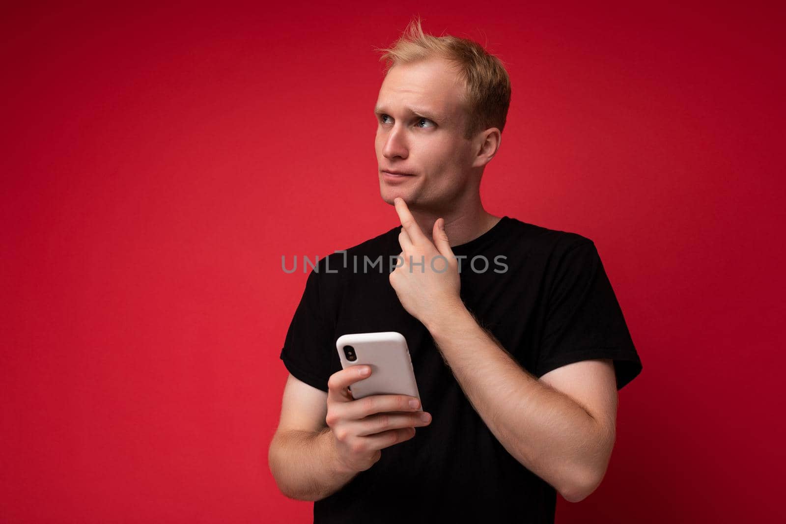 Thoughtful Handsome young man isolated over background wall wearing everyday clothes holding and using mobile phone writing sms looking to the side.