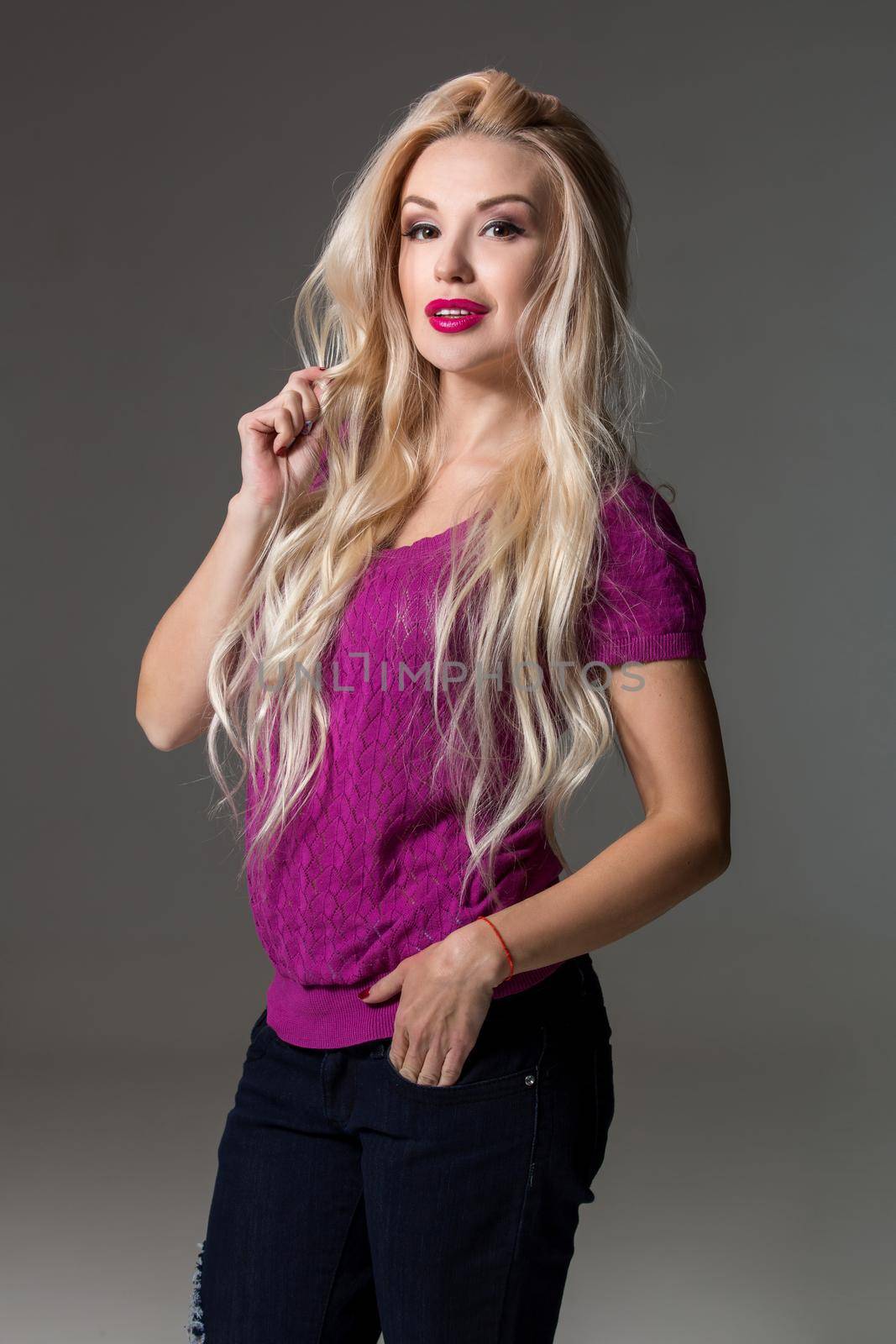 Happy young blond hair woman looking into the camera while standing against grey background. Young woman in a lilac blouse