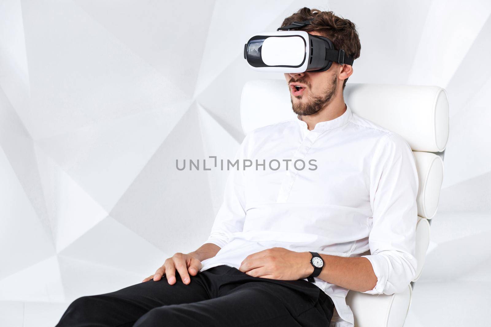 Excited young man is getting experience using VR-headset glasses of virtual reality gesticulating with his hands. A young man sits on a comfortable armchair in a room with white walls
