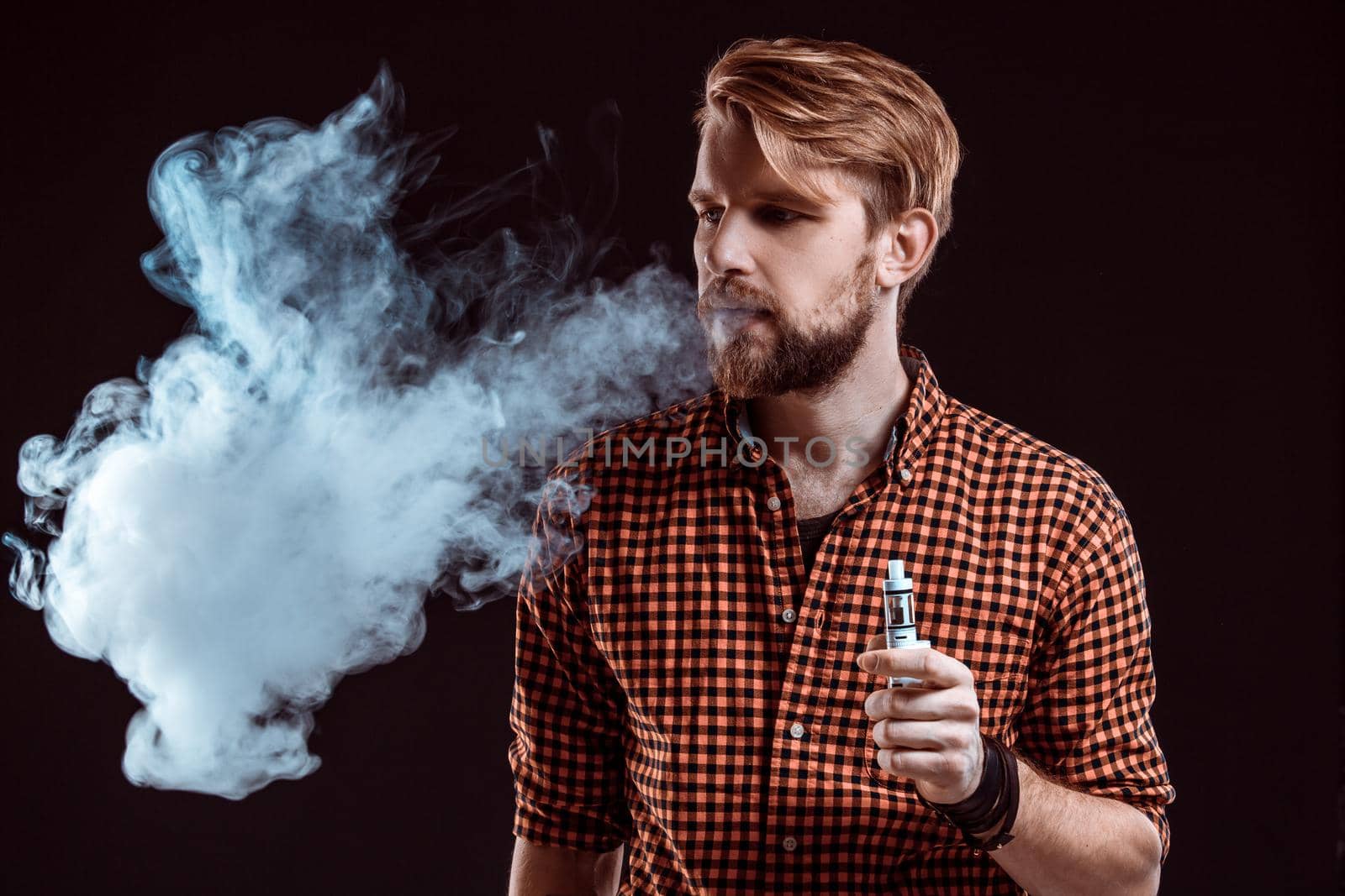 young man wearing a plaid shirt smokes an electronic cigarette on a black background