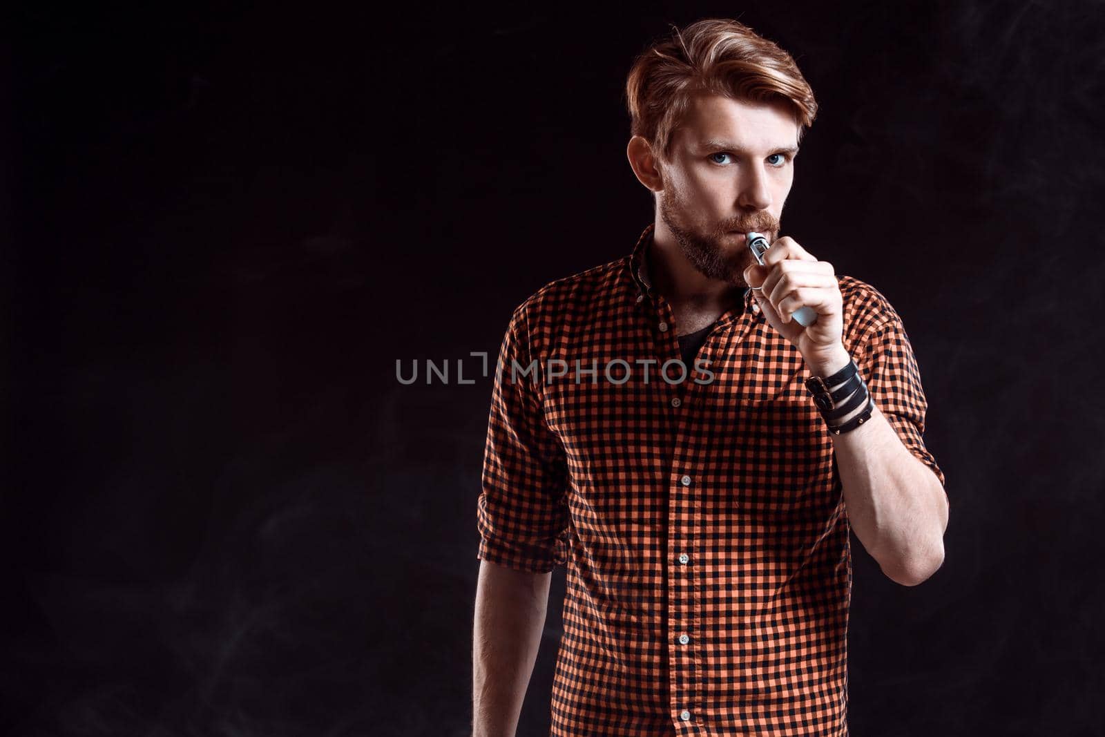 young man wearing a plaid shirt smokes an electronic cigarette on a black background. He looks into the camera